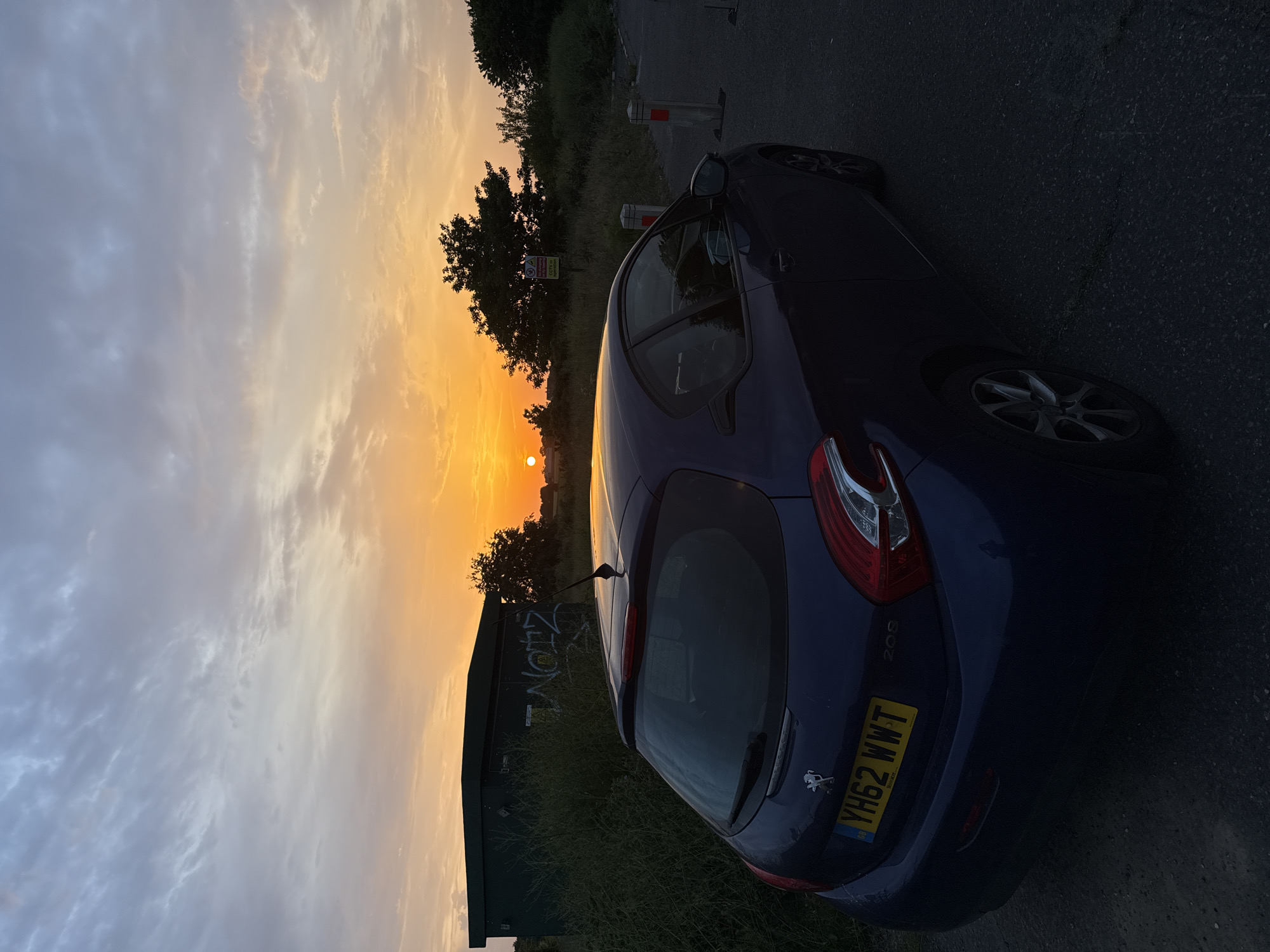 A parked blue Peugeot 208, license plate YH62 WWT, occupies the right side of the frame on a gray asphalt road. To the left of the car, a dark green electrical box displays graffiti. Beyond the box, trees and a setting sun paint the sky with orange and yellow hues. The location appears to be in Great Yarmouth, United Kingdom, based on the license plate. The sun is low on the horizon, near a line of trees. The overall environment has a rural feel.