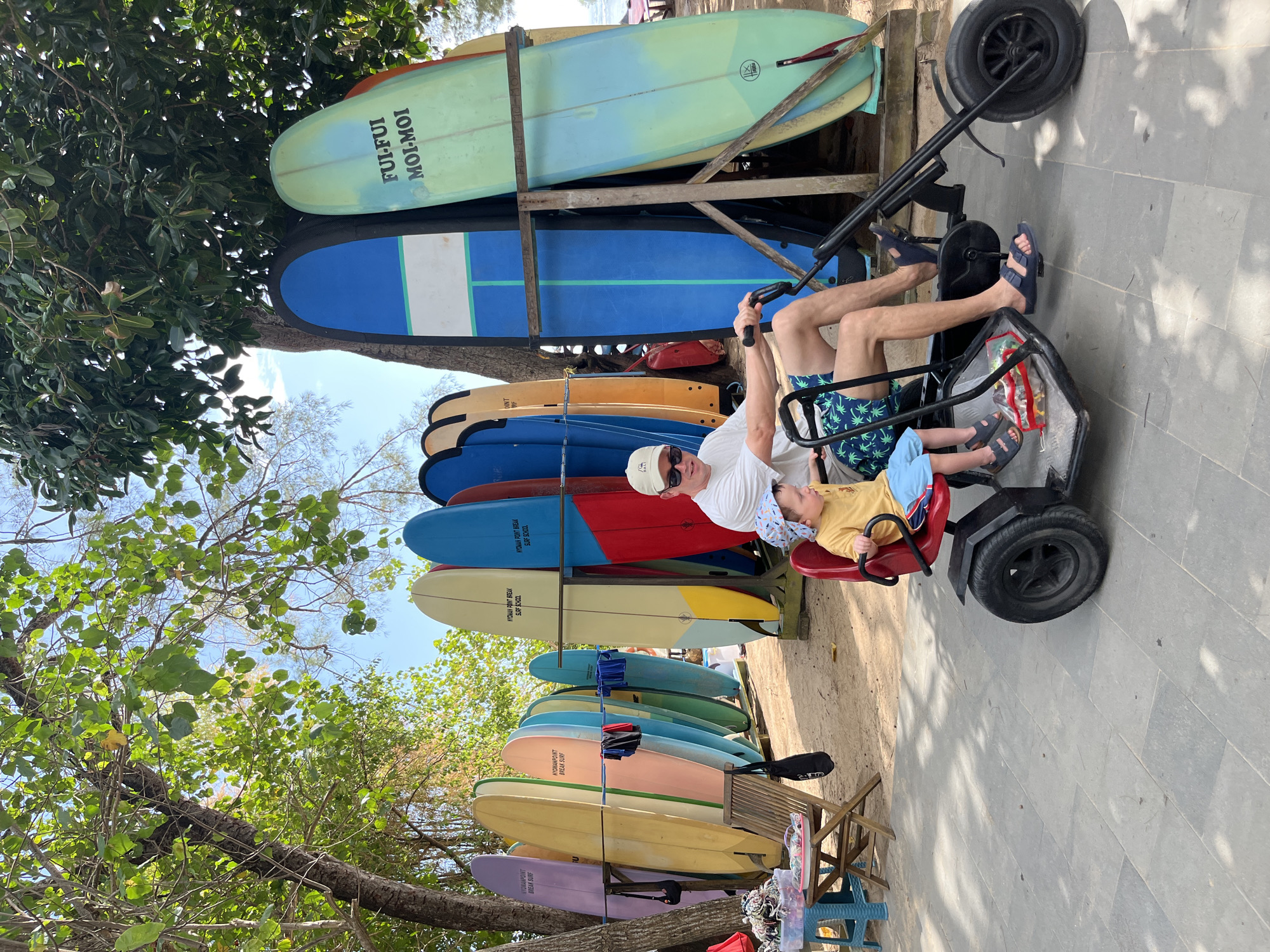 In Kuta Selatan, Indonesia, a man and a young child are on a black pedal-powered cart parked on a paved walkway. The man, wearing a white t-shirt, sunglasses, a white baseball cap, and blue patterned shorts, is at the controls of the cart. The child, in a yellow t-shirt and blue shorts, is seated in front of him. Behind them, a rack holds a collection of colorful surfboards of various sizes and designs, suggesting a beach or surf school location. The environment appears sunny, with trees overhead casting dappled shadows on the ground. The scene is set outdoors, with sandy ground visible in the background.