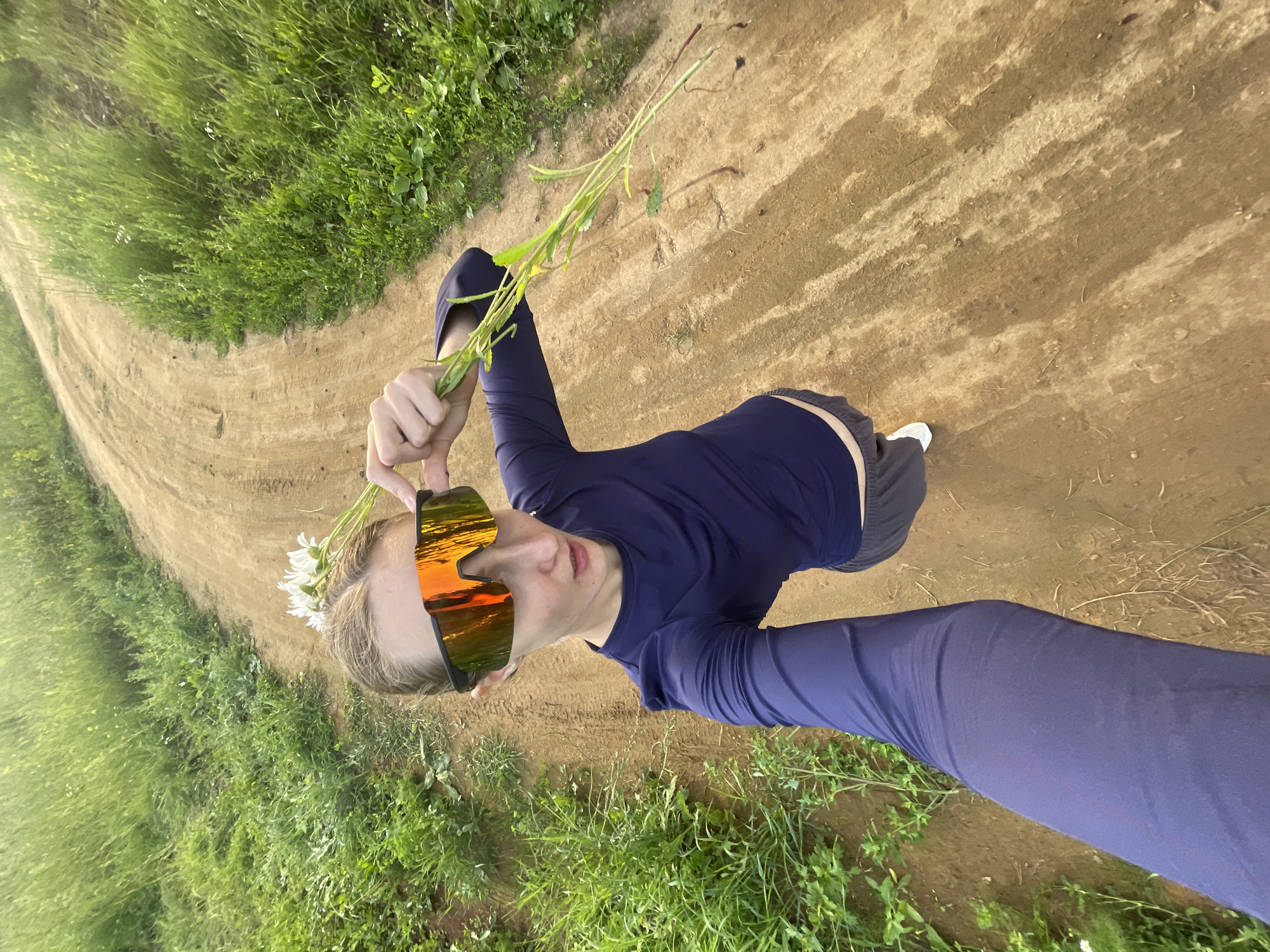 A person is holding a bouquet of wildflowers and looking at the camera. They are wearing a dark blue long-sleeved shirt, grey shorts, and white sneakers. Their sunglasses have a reflective orange and yellow tint, possibly from a sunset. The person is standing on a dirt path with green grass on either side. The scene appears to be outdoors in a rural area, likely in Russia, given the location context. The lighting suggests it is either early morning or late afternoon. The person's pose and direct gaze indicate they are posing for a selfie.