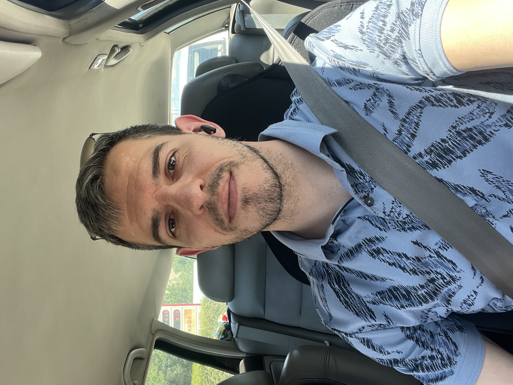 A man is in the driver's seat of a car, wearing a light blue collared shirt with a dark abstract pattern and a grey seatbelt. He has short, graying brown hair, a beard, and is wearing a black wireless earbud in his right ear. Sunglasses are perched on his head. The car's interior is visible, with grey seats and the car's headliner. Outside the car, through the windows, a bright sky and green trees can be seen, along with a red and yellow sign that appears to be a gas station price board with Cyrillic text "кафе" (cafe). The scene is likely a street in Sofia, Bulgaria, given the Cyrillic text.