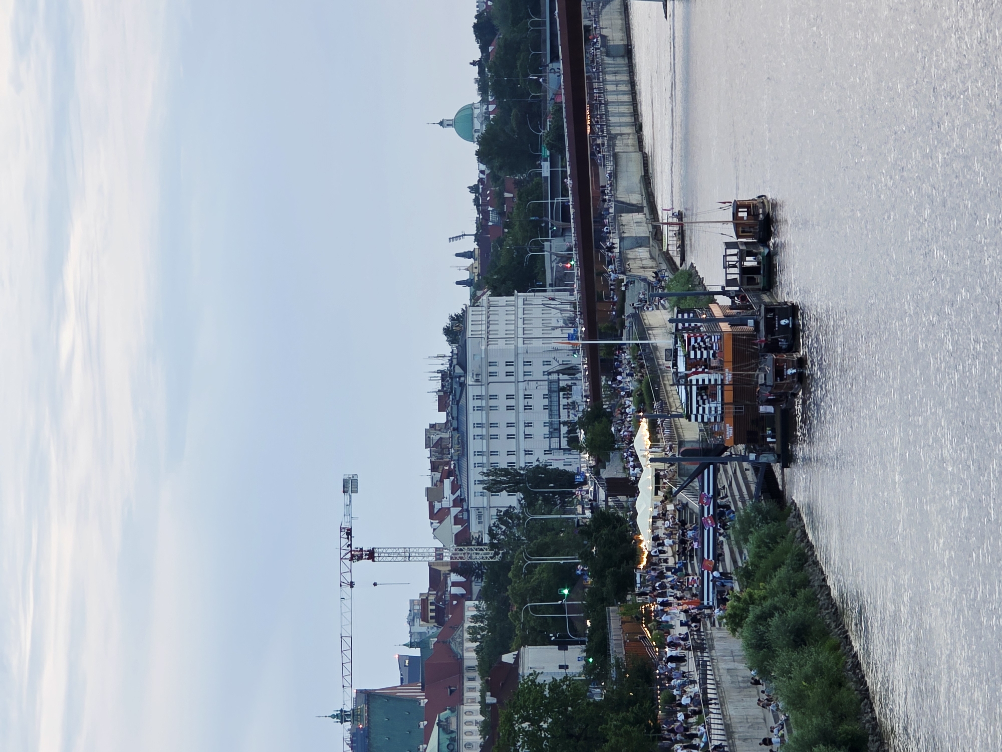 The image shows a view of the Vistula River in Warsaw, Poland, with the city's skyline in the background. On the riverbank, there is a promenade filled with people enjoying an evening outing. Several boats are docked along the river, with some appearing to be cafes or bars. A large red bridge spans the river in the distance. Buildings of various architectural styles, including a green-domed structure, are visible behind the promenade and trees. A construction crane is also present in the cityscape, indicating ongoing development. The sky is overcast, suggesting twilight or an early evening scene.