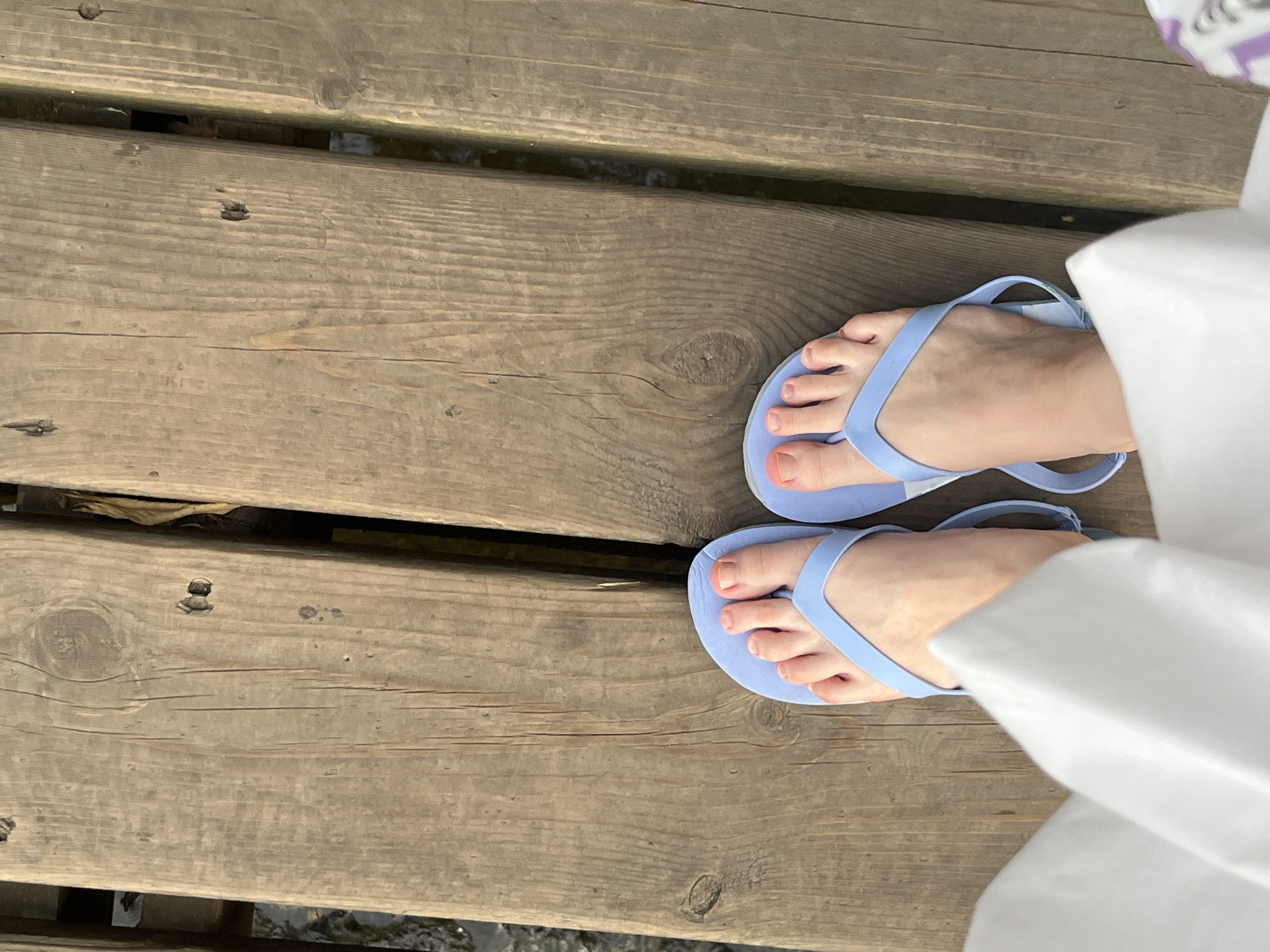A high-angle, medium shot captures a person's feet wearing light blue flip-flops standing on a wooden boardwalk. The boardwalk planks are oriented vertically and appear weathered. The person is wearing a white garment that partially covers their legs and feet. The scene is devoid of any specific landmarks indicating the city of Suzdal, Russia.