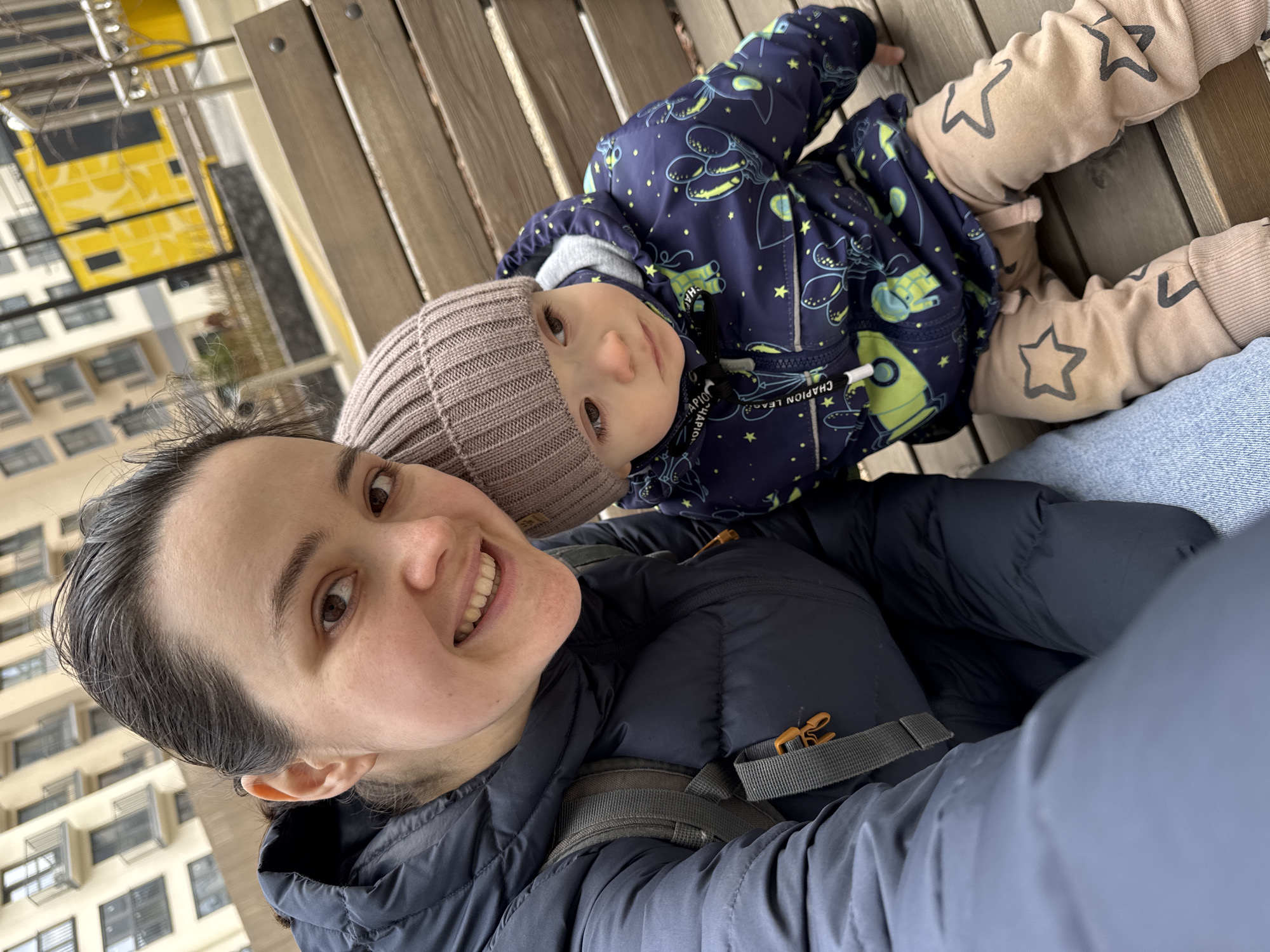A woman takes a selfie while lying on a wooden park bench in Izhevsk, Russia, with a young child lying on her stomach. The woman has dark hair and is smiling at the camera. She is wearing a dark-colored puffer jacket with a gray backpack strap visible. The child is wearing a brown knit hat, a blue jacket with rocket ship patterns, and beige pants with star designs. In the background are buildings and a yellow structure with black markings. The image is rotated slightly to the left.