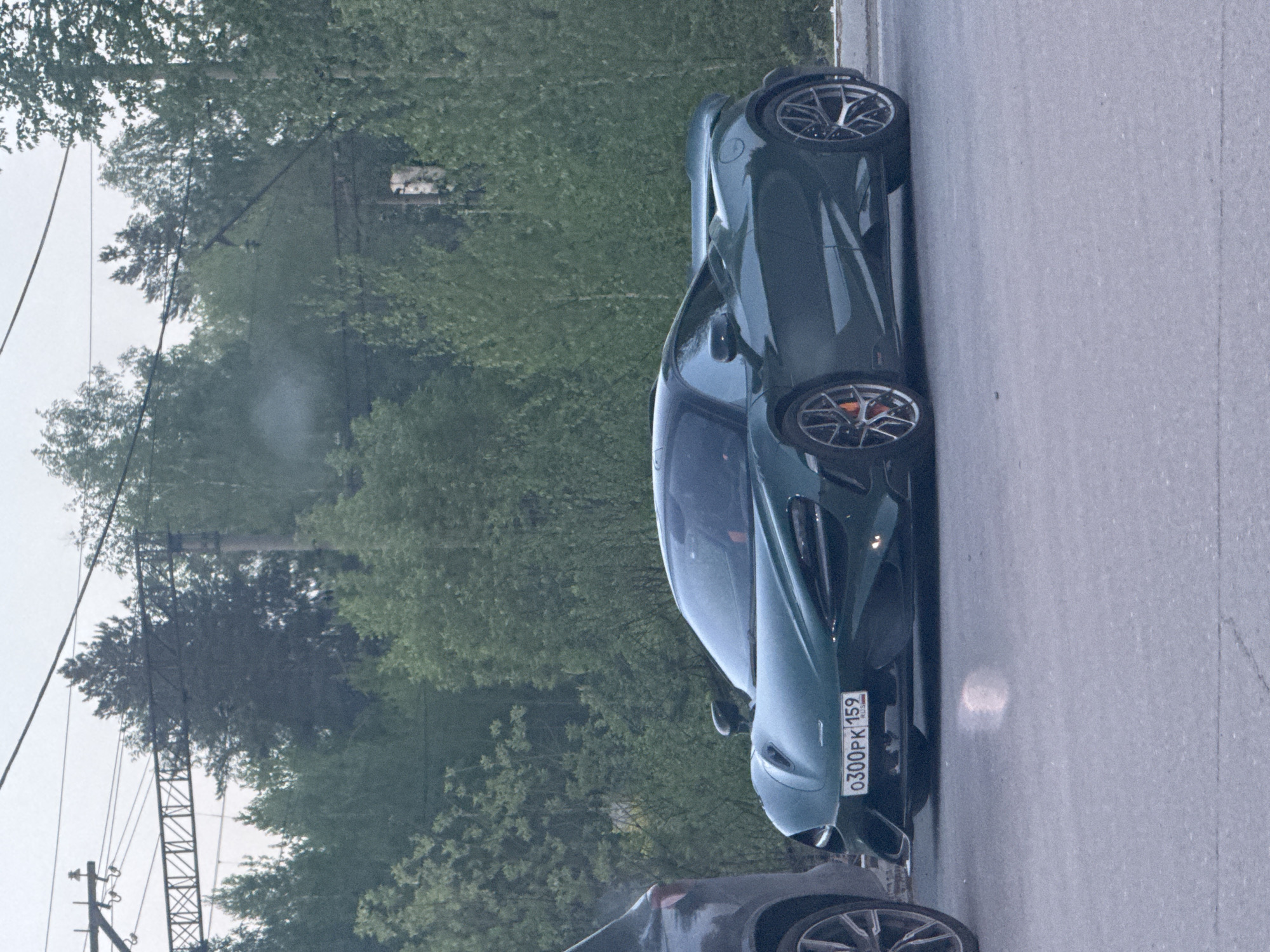A green McLaren sports car is parked on a street in Perm, Russia. The license plate reads "O300PK 159 RUS". A section of another car is visible at the bottom of the image. Trees and utility poles line the side of the road, suggesting a location on the city's edge. The weather appears overcast.