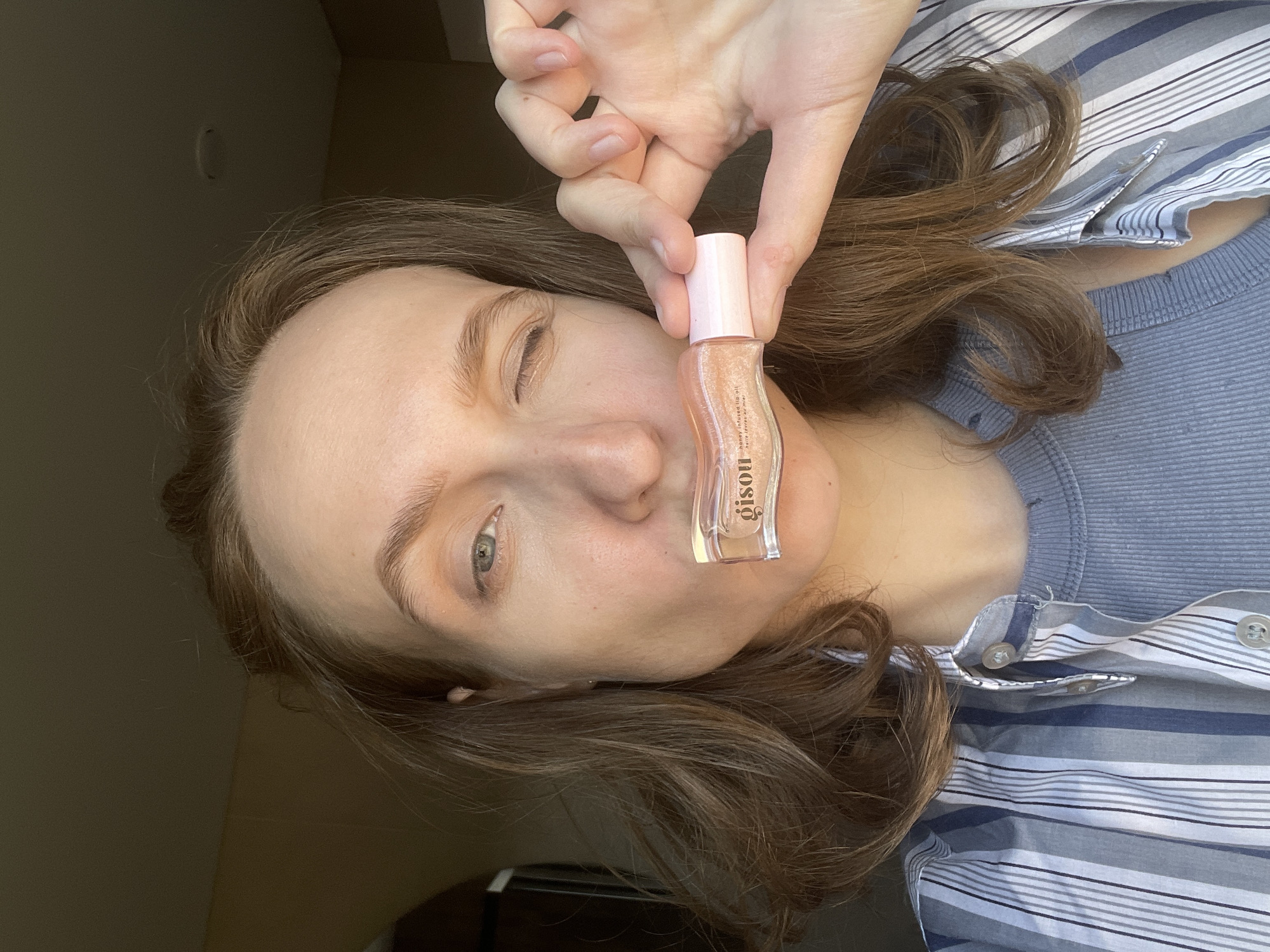 A woman is holding a small, pink-capped bottle of Gisou Honey Infused Lip Oil in front of her face, partially obscuring her left eye. She has fair skin, light brown hair, and her left eye is visible. She is wearing a light blue top and a striped shirt, and the background appears to be indoors. The location in the city of Подшивалово, Russia, is not determinable from the image.