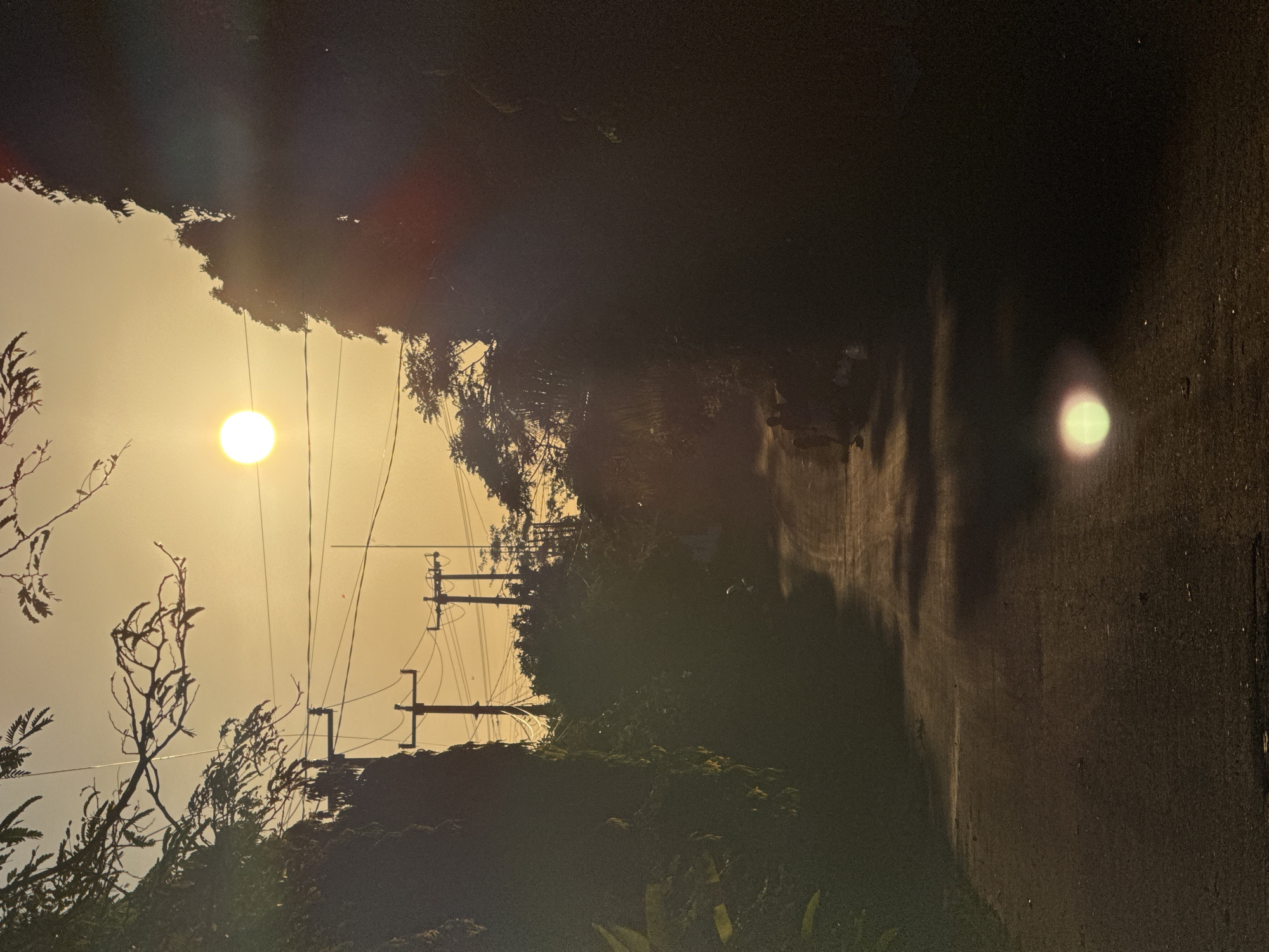 The image captures a view along a road in Ungasan, Indonesia, during a sunset. The sun is visible, casting a strong light across the landscape. The road, slightly curved, stretches into the distance, flanked by trees and vegetation. Utility poles and electrical wires are positioned along the road, with shadows extending across the pavement.
