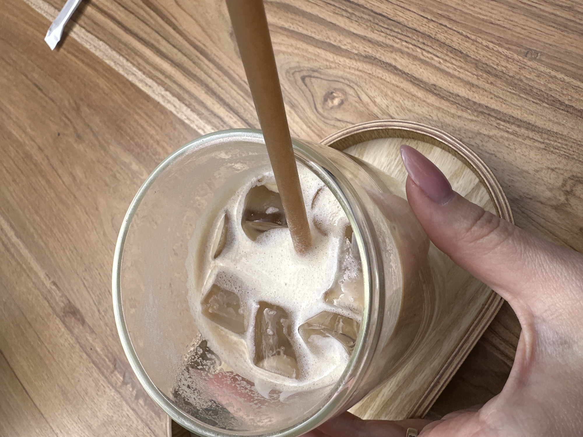A high-angle shot reveals a hand holding a glass of iced coffee on a wooden coaster, set upon a wooden table. The coffee, mixed with ice cubes, exhibits a foamy layer on top, and is being stirred with a brown straw. A portion of a white sugar packet lies on the table's surface. The scene lacks identifiable landmarks, precluding specific location within Tbilisi, Georgia. The focus is primarily on the iced coffee and the action of holding it.