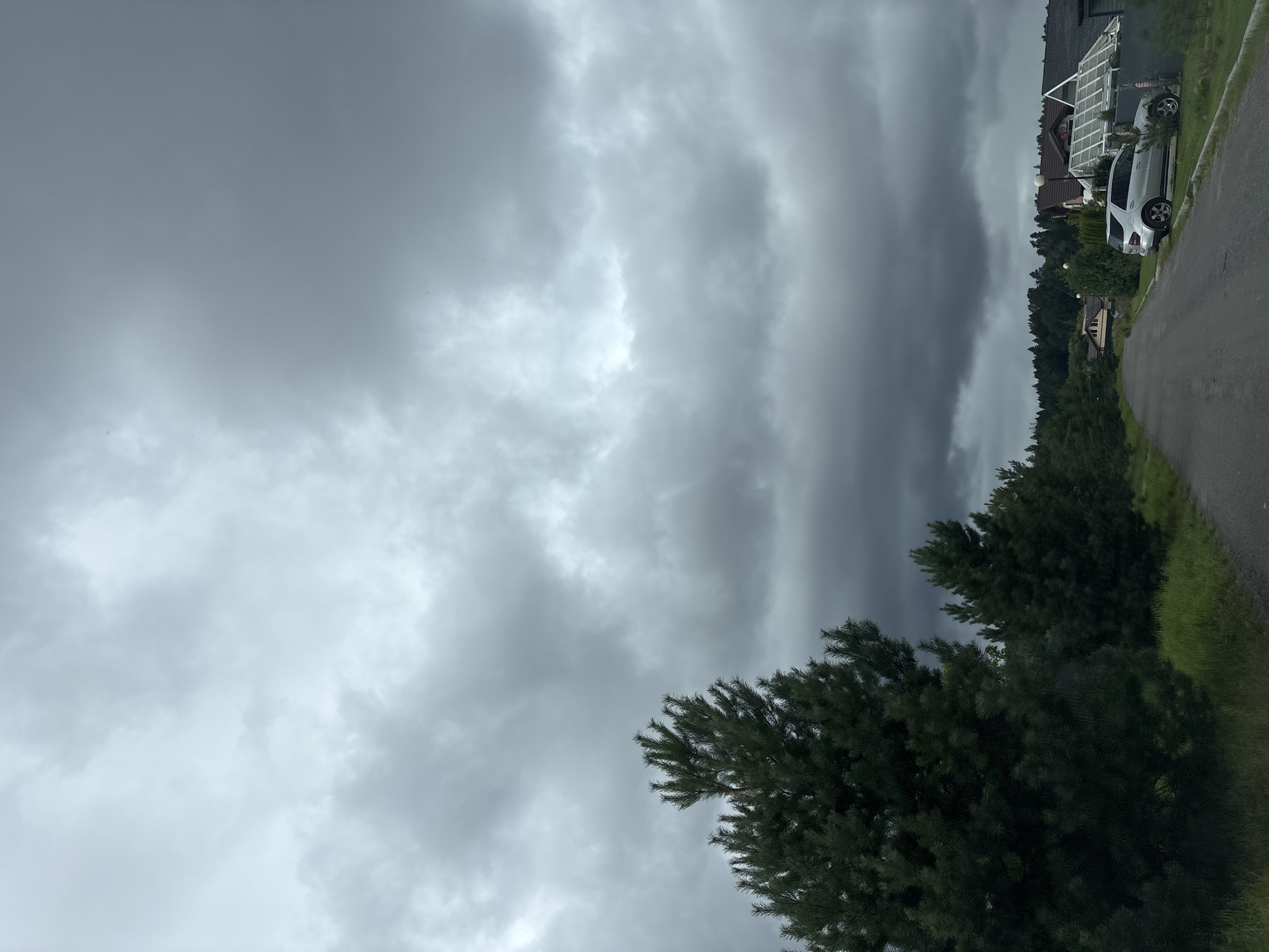The image depicts a suburban street in Zvenigorod, Russia, under a heavily overcast sky. The road, paved with asphalt, winds through a sparsely populated area with a line of evergreen trees on the left. On the right side of the road, several houses with visible roofs and architectural details are visible, along with a silver SUV parked on the grass next to a driveway. The sky is filled with dark, heavy clouds, suggesting an impending storm or recent rainfall. The overall atmosphere is somber and subdued due to the weather conditions.