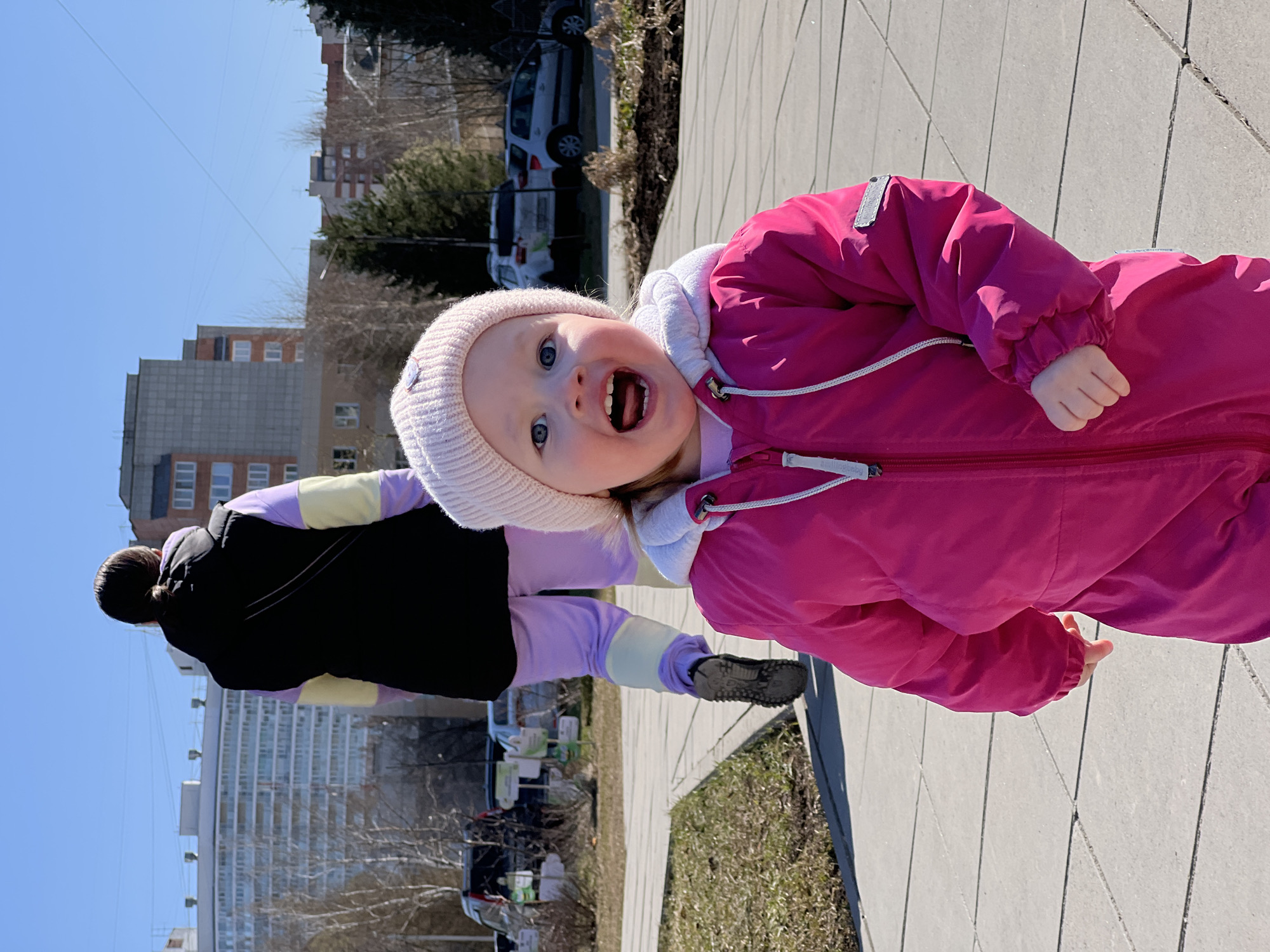 A fair-skinned child wearing a pink snowsuit and a light pink knit hat is the focus of this outdoor photo. The child is facing the camera, mouth open in what appears to be a shout or wide smile. The background includes a sidewalk, a strip of grass, and several multi-story buildings, which suggest a residential area. A person in a black, lilac, and yellow jacket is walking near the child, blurred in the background. The photo was taken in the city of Perm, Russia.