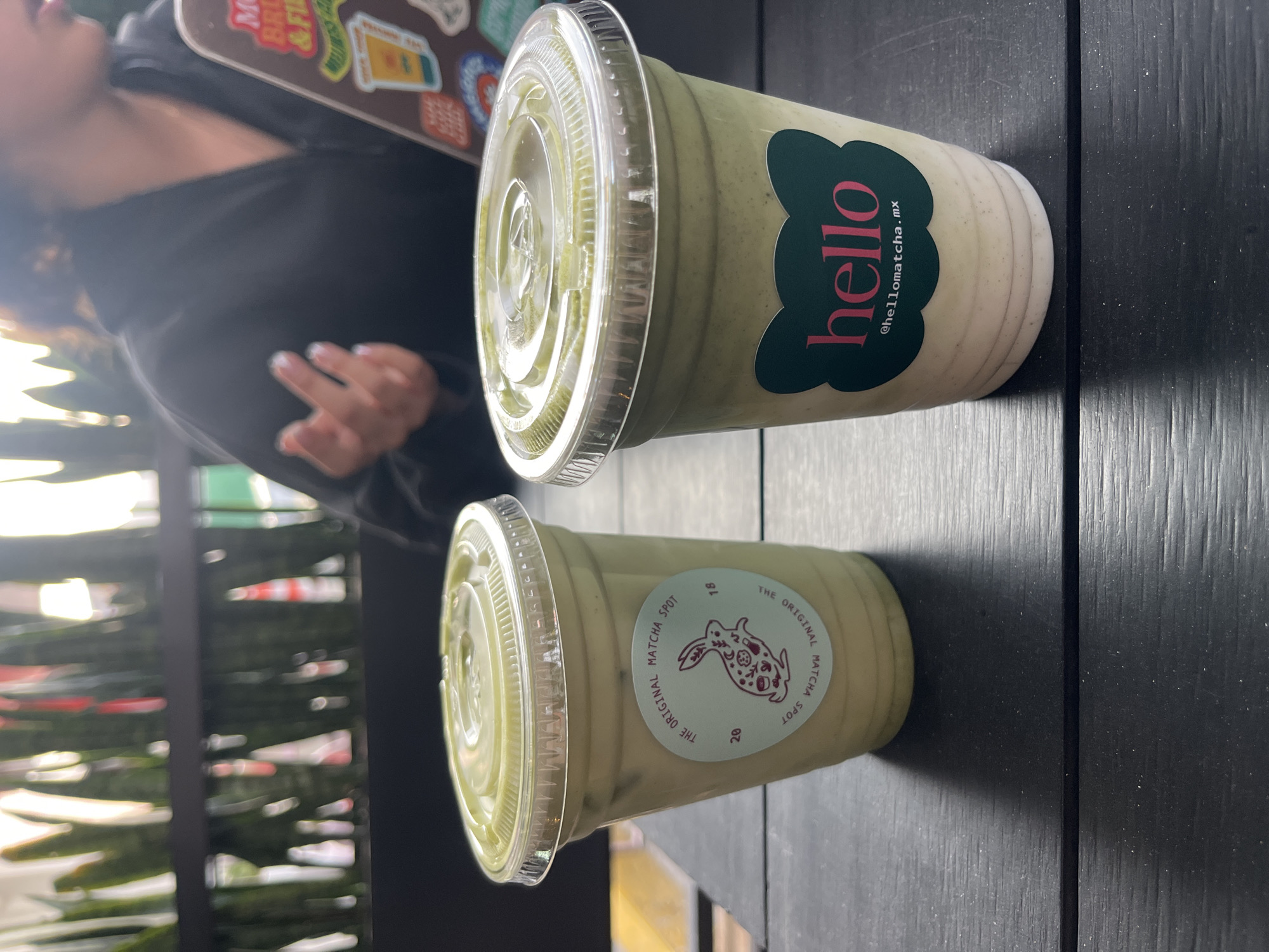 The image shows two plastic cups of matcha drinks sitting on a dark, possibly wooden, surface. The cup closer to the camera has a sticker on it that reads "THE ORIGINAL MATCHA SPOT" in a circular pattern around a bunny design. The other cup has a logo that reads "hello" in pink, with "@hellomatcha.mx" underneath. A person is partially visible in the background wearing a black shirt. Based on the logo "@hellomatcha.mx" which suggests this is in Mexico, and the location identified in the prompt, the image was taken in San Bernardino Tlaxcalancingo, Mexico.