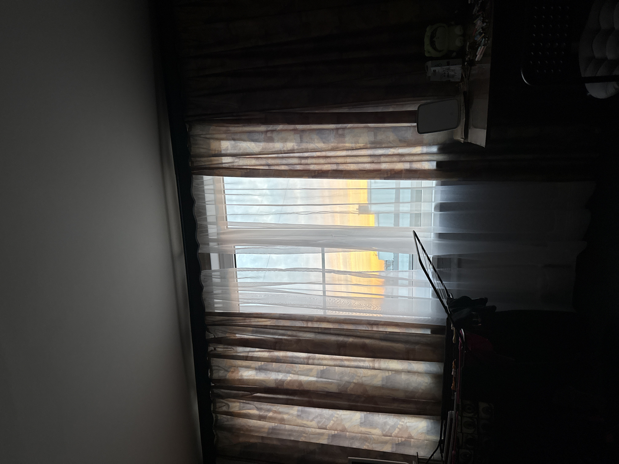 A window in a room is visible, covered with curtains and white blinds. Light streams in through the blinds, suggesting the presence of sunshine or daylight outside. The window faces a structure with a pale, yellow-tinted facade. Due to the distinct architecture and color, the structure may be St. Isaac's Cathedral in Saint Petersburg, Russia. Part of a dark object and some miscellaneous objects are positioned by the window. The room itself is dimly lit, with the left wall a pale gray.