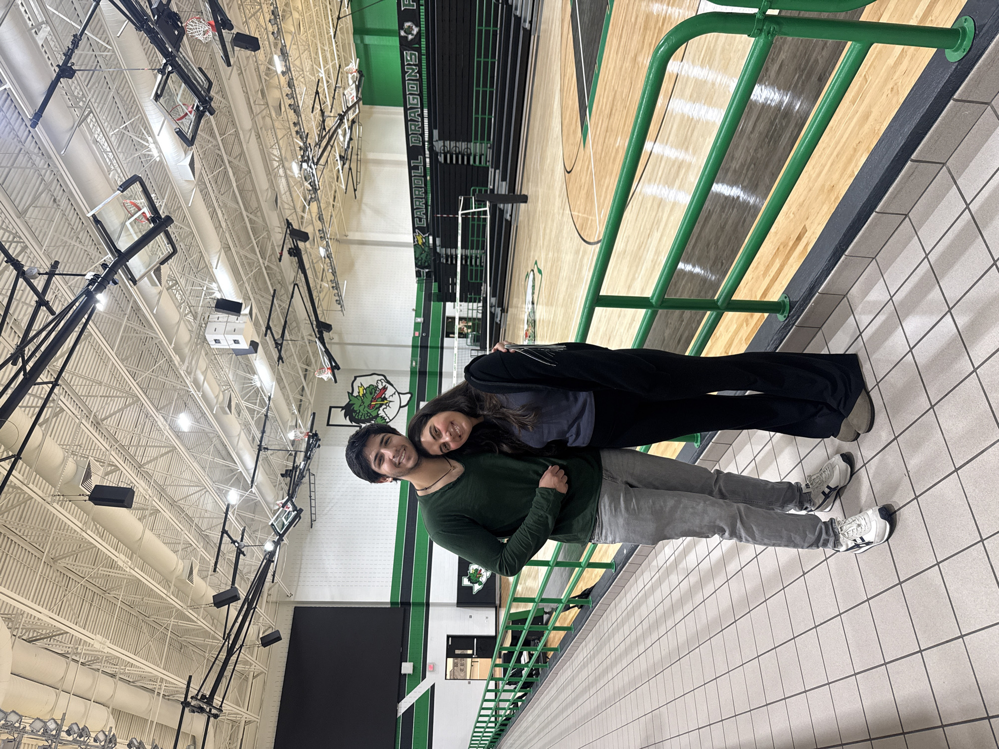 Two individuals are standing in the Carroll Senior High School gymnasium, located in Southlake, Texas, United States. The male is wearing a green sweater and grey jeans with white sneakers, while the female is wearing a dark top and black pants with light-colored sneakers. They are posing with the female leaning her head on the male's shoulder. The gymnasium features basketball courts, retractable bleachers with the school's "Carroll Dragons" logo, and overhead lighting and ventilation systems. The floor is tiled with white and grey squares, and there's a green railing behind them.