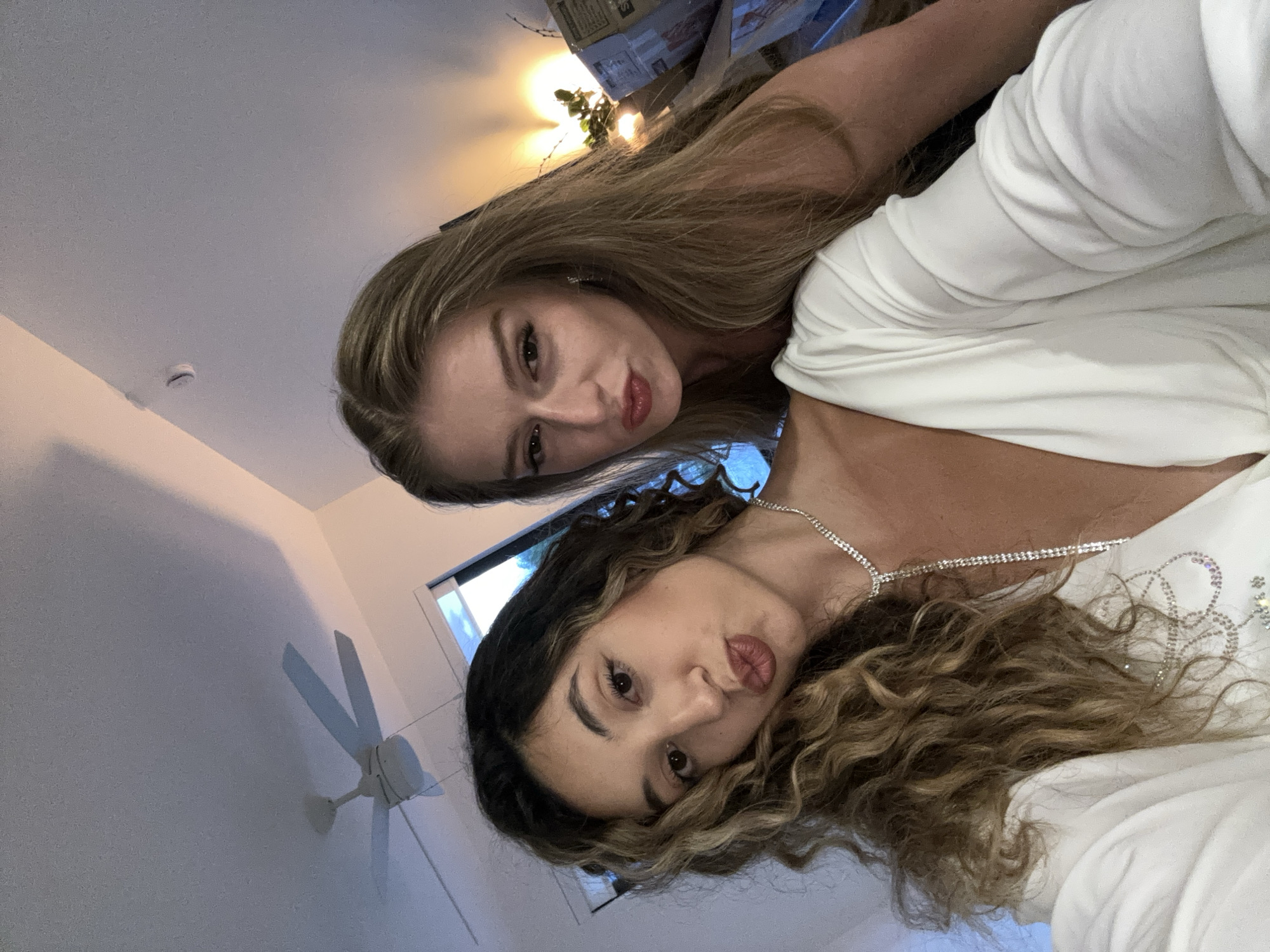 Two young women are captured in a selfie taken indoors, likely in Hallandale Beach, Florida, United States. The woman in the foreground, on the left, has long, curly, dark brown hair with blonde highlights. She is wearing a white top with a low-cut neckline and a sparkling necklace. Her lips are pursed in a kiss pose. To her right, the second woman has long, straight, blonde hair. She is also wearing a low-cut white garment and has her lips pursed. The background shows a ceiling fan, a window with a view of the outdoors, and stacked boxes, suggesting a casual setting, perhaps a home. The lighting is warm and slightly dim, with artificial lights visible in the background.