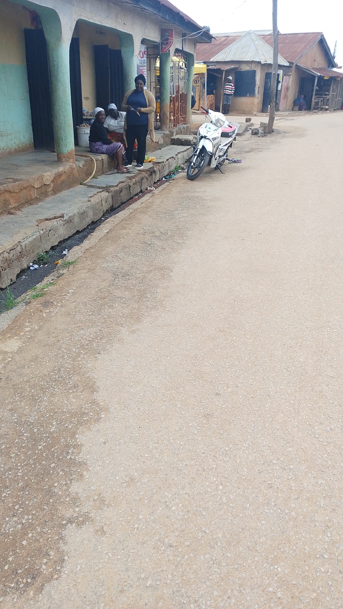The image depicts a street scene in a possibly rural or semi-urban area. In the foreground, a paved road shows signs of wear and tear, with patches of lighter and darker asphalt. Along the left side of the road is a raised concrete drainage channel, filled with debris and some sparse green vegetation.
Adjacent to the drainage channel are buildings with open doorways and awnings. In front of one of these buildings, three people are gathered. Two individuals, appearing to be elderly women wearing headscarves, are seated on a low concrete step. One is wearing a purple patterned skirt and a dark top, while the other is wearing a white headscarf and a light-colored top. Standing next to them is a third person, likely a woman, dressed in dark pants and a beige cardigan, holding a dark object, possibly a mobile phone, in her hand.
Further down the street, a white motorcycle is parked on the right side of the road, facing towards the viewer. Beyond the motorcycle, more buildings with corrugated metal roofs are visible, suggesting a commercial or residential area. Another person in a striped shirt can be seen standing near one of these buildings. The overall environment appears to be dry and dusty, consistent with a tropical or subtropical climate. The lighting suggests it is daytime, with overcast or diffused sunlight.