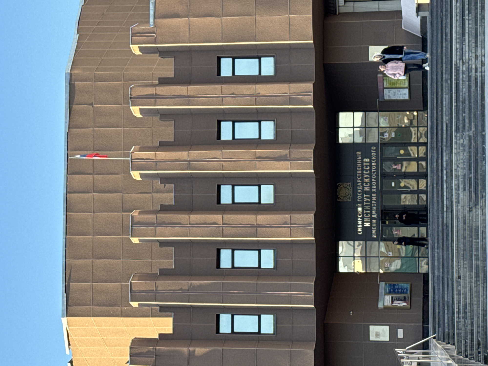 The image shows a building facade in Krasnoyarsk, Russia. The structure is brown, rectangularly sectioned, and punctuated by rows of rectangular windows. A flag is visible towards the top left. To the right of the building's facade is a large entrance with glass doors, and signage written in Russian. The sign indicates the building is the Siberian State Institute of Arts named after Dmitri Hvorostovsky. A pair of individuals are standing on the stairs leading up to the entrance. Other people can be seen entering and exiting the building. The weather appears to be clear and sunny.