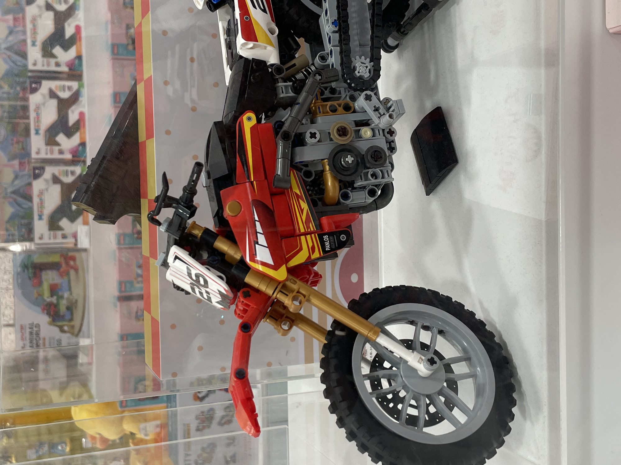 The image shows a close-up view of a red and gold Lego Technic model motorcycle displayed in a retail setting. The motorcycle is detailed, featuring a front suspension, spoked wheels with rubber tires, a chain drive mechanism, and racing decals. The number "25" is visible on a white plate at the front. The motorcycle is positioned on a white surface, likely a display shelf, with a clear plastic barrier in the foreground. In the background, several boxes with colorful graphics, possibly toys or games, are visible, along with a yellow stuffed toy peeking from behind a vertical support. The lighting suggests an indoor environment, consistent with a store or exhibition. The image does not provide any information or visual cues that would allow for the determination of its location within Davao City, Philippines.