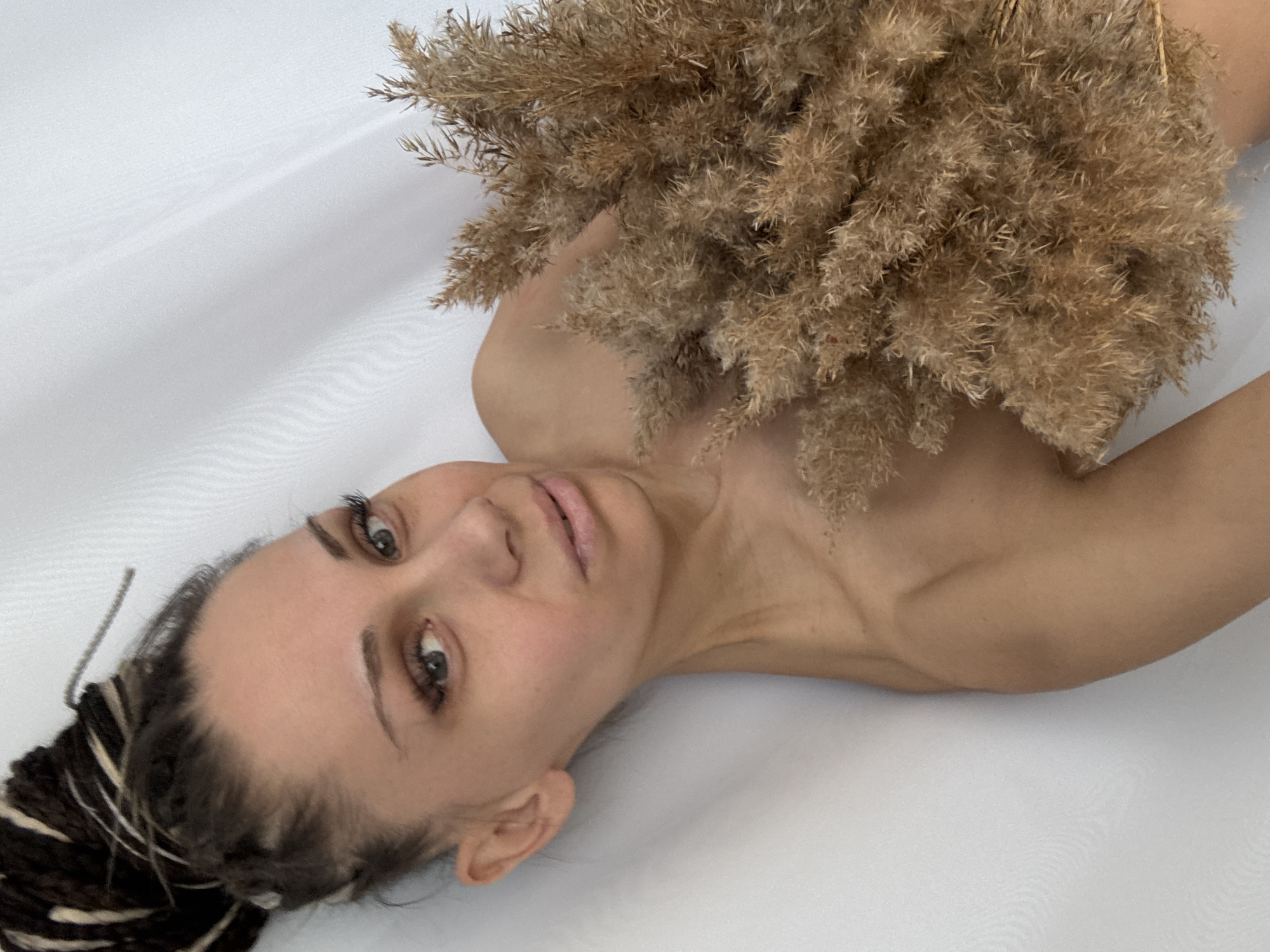 A woman with braided hair lies on a white surface. She is holding a bunch of dried grass or wheat stalks over her chest. Her head is tilted towards the left, and she is looking towards the camera. The background is plain white, and the setting appears to be indoors. There is no indication of the city of Rusanovo or any other location.