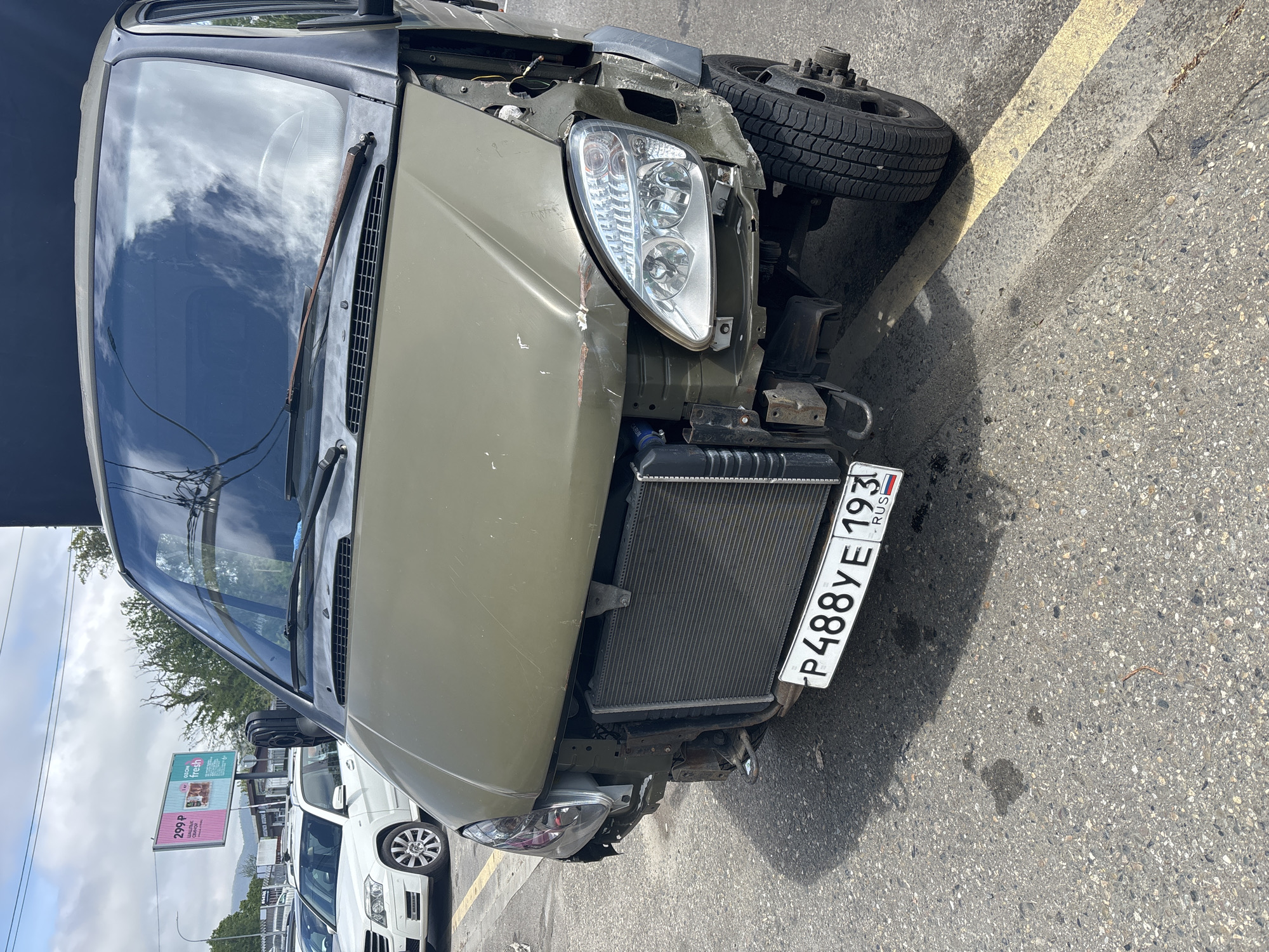 The image shows the front of a damaged, olive-green van, missing its bumper and with a visible radiator. The van's license plate reads "P488YE 193 RUS," indicating a Russian vehicle registration. The front left headlight is still intact, though surrounded by damaged bodywork. The vehicle appears to be parked on asphalt, next to a yellow painted line. In the background, a white SUV is visible, along with a billboard advertising a deal for "299P" for something that is "fresh." Based on the license plate and the text on the billboard being in Russian, the location is in Russia. Given the "193" region code on the license plate, it is likely in the city of Sochi.