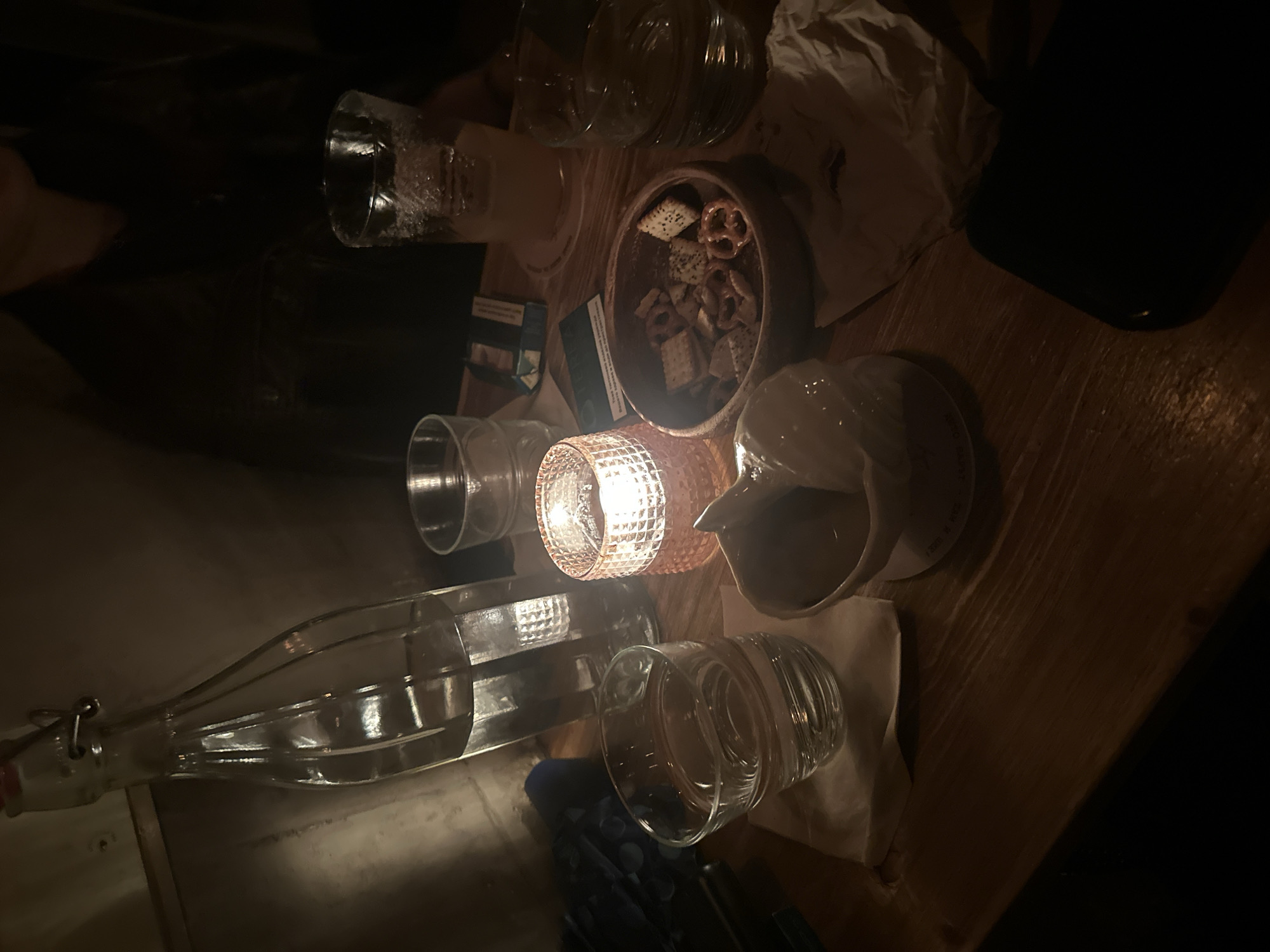 The image shows a dimly lit scene on a wooden table in a bar or restaurant in Varna, Bulgaria. There's a mixture of empty drinking glasses, a glass water bottle, a small dish containing pretzels and crackers, a small pitcher, a pack of cigarettes, some crumpled napkins, and a candle holder with a lit candle. A dark object, likely a phone or wallet, is on the right edge of the table. The lighting is soft and indirect, originating from the candle and potentially other sources outside the frame. The focus is sharpest on the items clustered around the candle. The setting appears relaxed and informal. There are no people fully visible in the frame, though a portion of an arm is barely visible in the upper left.