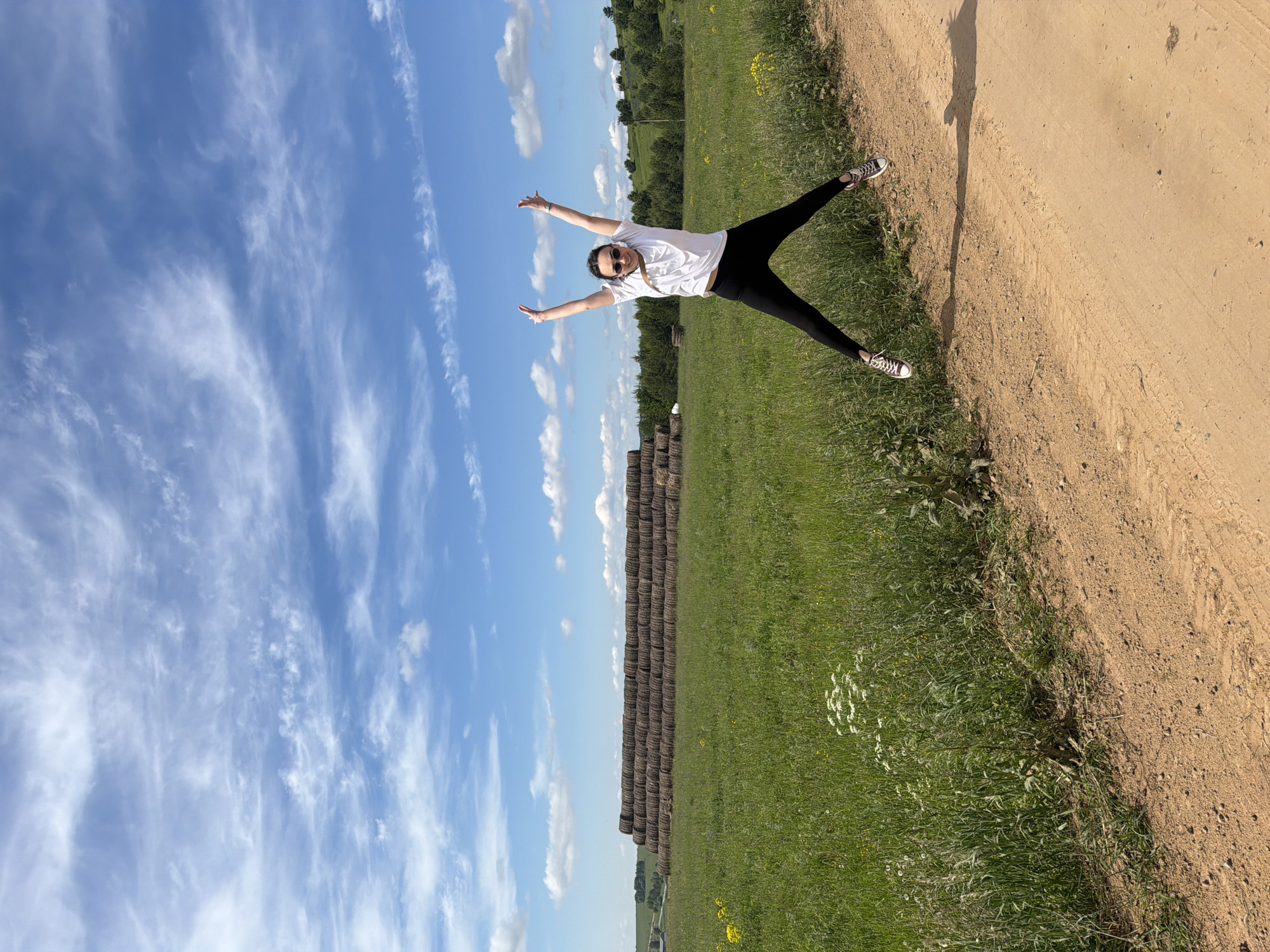 This vibrant image, taken in Van'ki, Russia, captures a person striking a joyful pose in a sunlit rural landscape. The photograph is oriented sideways, with the subject appearing to stand on the edge of a dirt path.
The main subject is a person, likely female, dressed in a white t-shirt, black leggings, and black and white sneakers. They wear sunglasses and a bright smile, with their arms outstretched and legs spread wide in an energetic, star-like pose, conveying a sense of happiness and freedom. To their right, a long, faint shadow of a person is cast on the light brown dirt path.
The setting is an open countryside environment. Extending behind the person and into the background is a vast green field, interspersed with wild plants and some white and yellow flowers. A prominent feature in the mid-ground is a long, continuous line of round hay bales, stacked several high, stretching horizontally across the field. Distant green trees are visible along the horizon.
The sky above is a brilliant blue, dotted with scattered white, wispy clouds, indicative of clear and pleasant weather. The strong sunlight suggests it is daytime, possibly late morning or afternoon. The overall scene depicts a peaceful agricultural or natural area with a wide-open expanse. No visible text is present in the image.