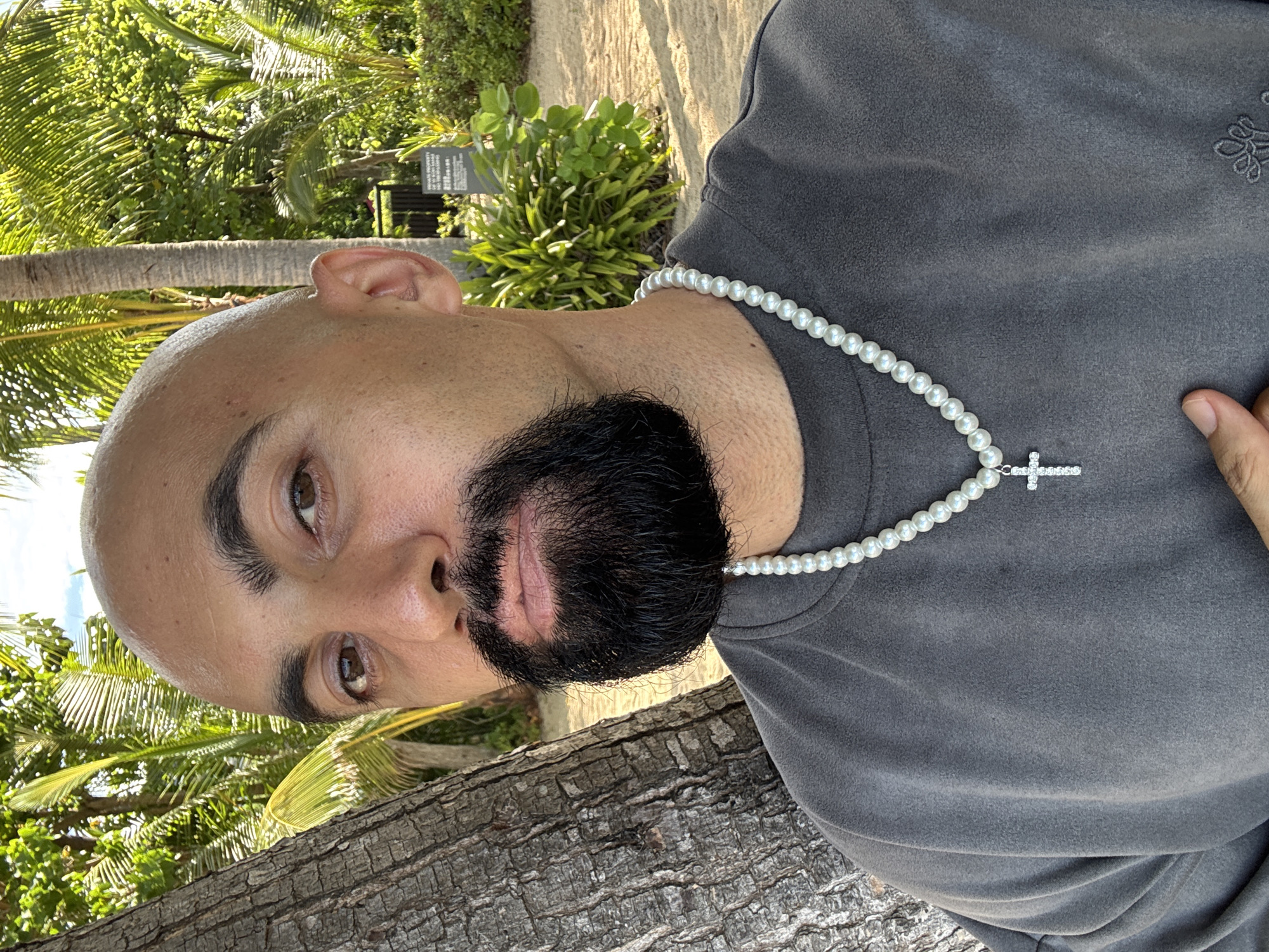 The image shows a close-up, waist-up portrait of a bald man with a full beard, wearing a grey sweatshirt and a pearl necklace with a cross pendant. He is positioned to the left of the frame, leaning against a tree trunk. The background features lush green palm trees and foliage, with sandy ground visible. A sign with text in Thai script is partially visible in the background, suggesting the location might be a resort or a public area in a tropical environment. The lighting suggests it is daytime, with sunlight casting shadows. The man's expression is neutral. There is no indication of the city of Ko Samui District, Thailand specifically, however, the tropical scenery is consistent with such a location.