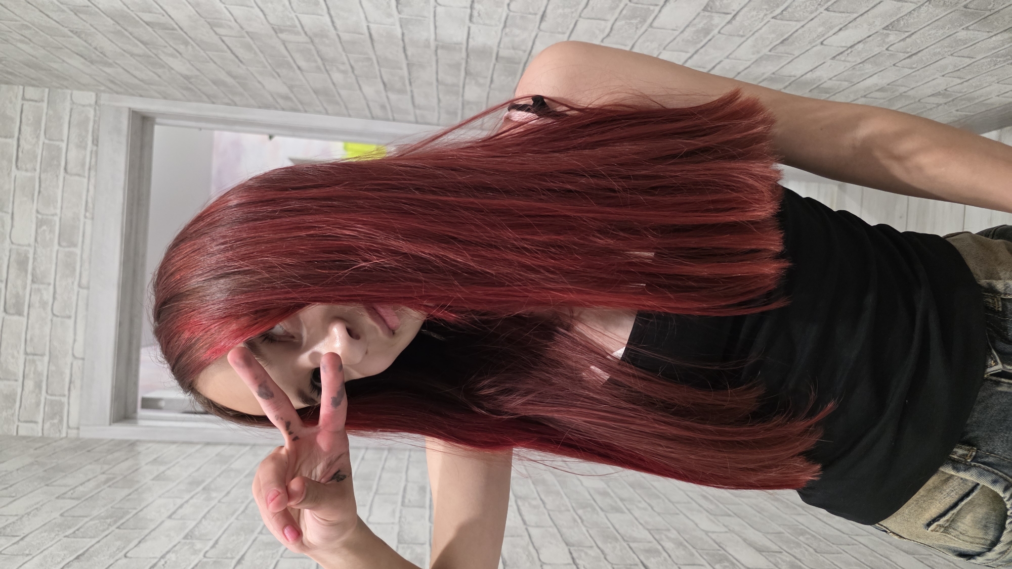 A young person with long, dark red hair is holding up a "peace" sign with their left hand, which has black smudges on the fingers. The person is wearing a black t-shirt and blue jeans. The background appears to be a room with white brick walls and a doorway with a white frame. The person's face is partially obscured by their hair. The image is oriented sideways. The location is a room with white brick walls and a doorway with a white frame, in the city of Unknown, Unknown.