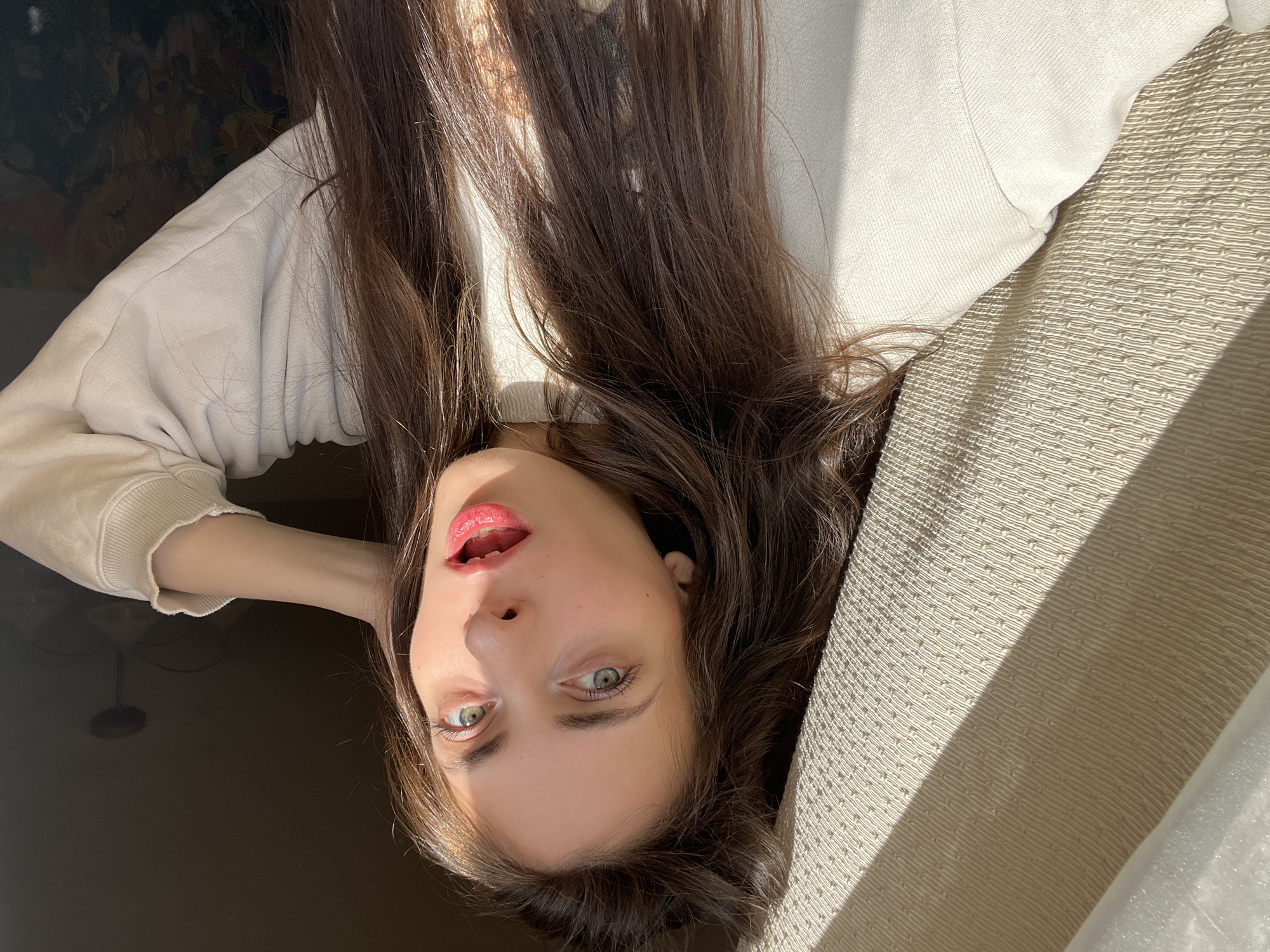 A fair-skinned woman with long brown hair lies on an upholstered, beige-colored object, possibly a sofa or lounge chair. She wears a light-colored sweater. Her head is tilted back, with her mouth slightly open and lips colored with a red gloss. Her eyes are green. The background features both dark and light areas, possibly indicating a room interior and a sunlit exterior. The photograph’s orientation suggests it was taken with the camera held upside down. It is not possible to confirm whether the location is Krasnoyarsk, Russia.