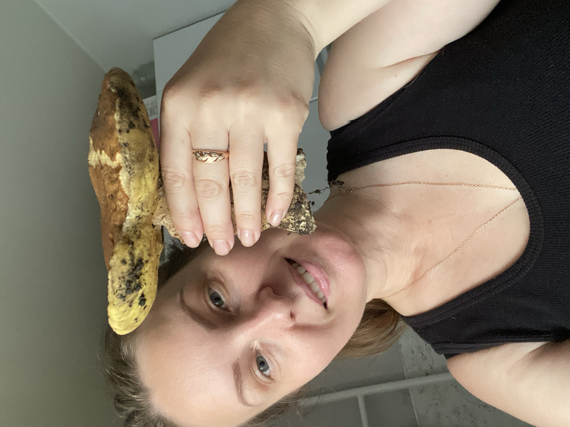A medium shot captures a woman with light skin and medium-length brown hair, pulled back, as she smiles and holds a mushroom close to her face. The mushroom has a tan and yellow cap, speckled with dark spots, and a thick stem with visible roots. The woman is wearing a black tank top and a delicate gold necklace. A thin gold ring with a leaf design adorns her left ring finger. In the background, a plain white surface is visible on the right, and a blurred, light-colored vertical object, possibly part of a shelf or furniture, is on the left. The overall environment appears to be indoors, with neutral lighting.