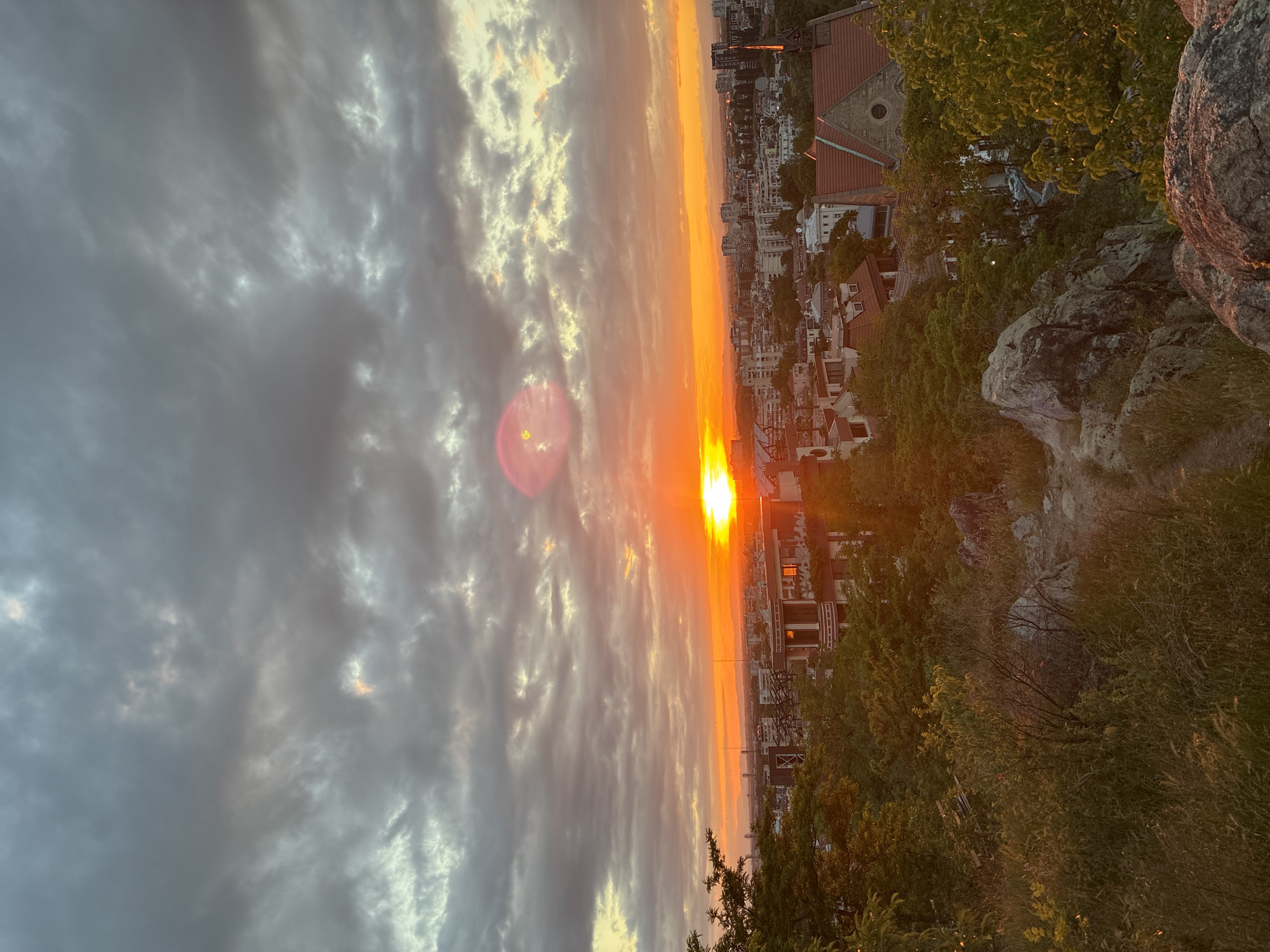 The image shows a sunset view over the city of Plovdiv, Bulgaria, likely taken from Nebet Tepe hill. The sun is setting on the horizon, casting a bright orange and yellow light across the sky and city. Clouds fill a significant portion of the sky, ranging from dark gray to light gray and white. Below the sunset and clouds, a cityscape is visible with buildings of varying heights and architectural styles. In the foreground, there are rocky formations and trees, suggesting the vantage point is a high, natural location. The image is rotated 90 degrees clockwise.