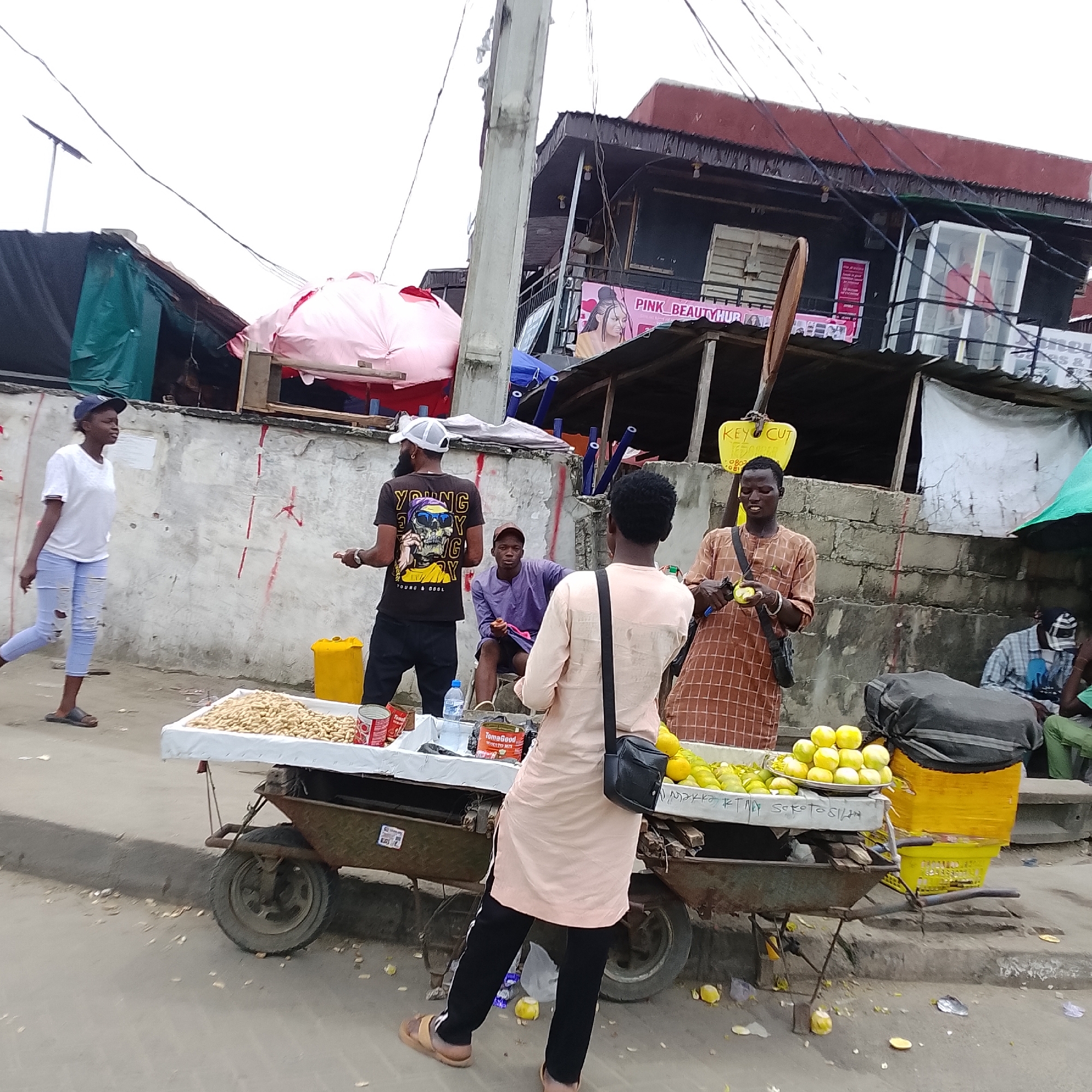 In Ado, Nigeria, a street scene unfolds with several individuals engaged in commerce and daily life. A young woman in a white t-shirt and ripped blue jeans walks past a stall. Beside her, a vendor in a black t-shirt with a graphic design displays canned goods and what appears to be peanuts on a cart. Another man, seated in a purple garment, observes the surroundings. A third man, in a patterned brown tunic, is actively engaged in preparing fruit, possibly limes or lemons, with a knife. In the background, buildings with signs such as "PINK BEAUTY HUB" suggest a commercial area. The environment is characterized by concrete walls, utility poles with overhead wires, and a general street setting with debris on the ground. The overall impression is a bustling local market or street-side business area.
