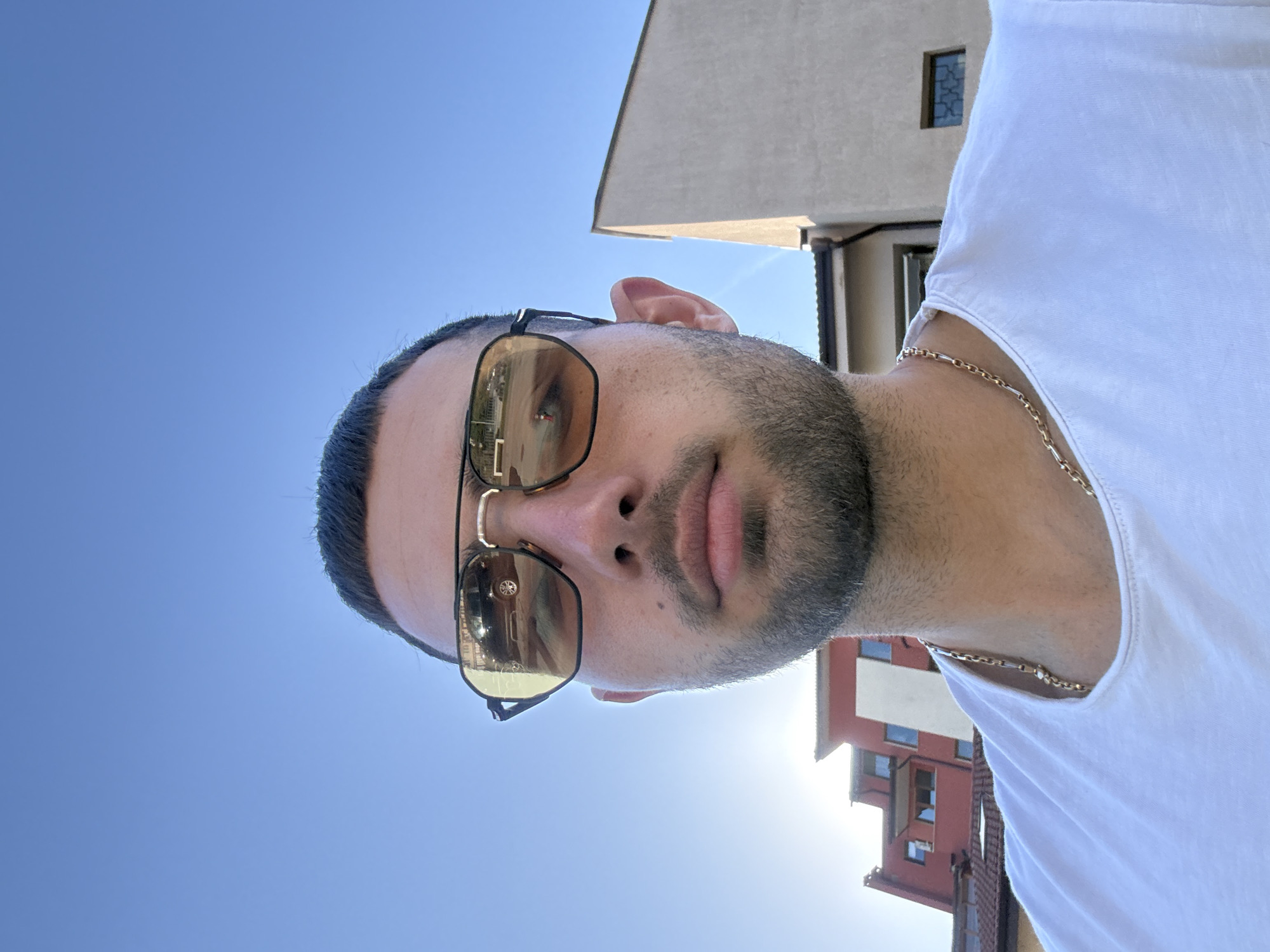A man wearing sunglasses and a white t-shirt stands outside. A gold chain is visible around his neck. The sky is blue, and buildings are in the background. The location appears to be Gabrovo, Bulgaria, based on the architecture.