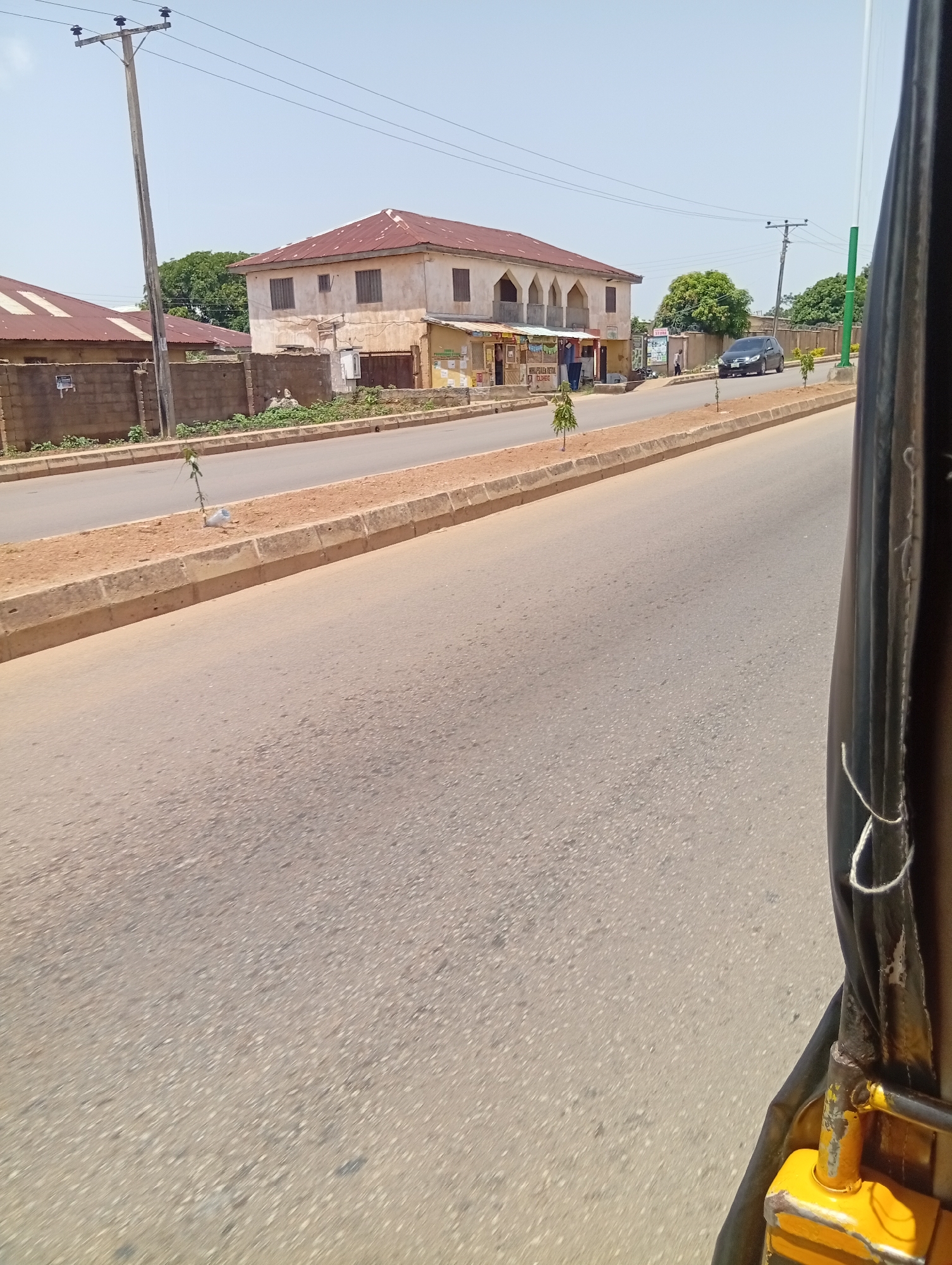This image depicts a street scene in Jos, Nigeria, under a bright, clear sky, suggesting midday. The foreground is dominated by a paved road, with the side of a yellow and black vehicle, possibly a tuk-tuk or auto-rickshaw, visible on the right. The vehicle's black fabric covering and yellow metal frame are prominent.
Across the road, a raised median with a concrete curb separates the lanes and is lined with small trees. Beyond the median, a two-story building with a weathered facade and a reddish-brown corrugated iron roof stands prominently. The building has arched openings on its upper level and what appear to be shutters on its windows. A storefront with signage is visible at the ground level. To the left of this building, another building with a similar roof is partially visible behind a brick wall.
Further down the street, more buildings and trees can be seen. A dark-colored car is parked on the side of the road. Utility poles with electrical wires are also present. The overall scene suggests a typical urban or semi-urban environment in Nigeria, with everyday life unfolding. There is no discernible emotion, but the bright sunlight and clear sky suggest a warm and dry day. Some text is visible on signs in front of the storefronts, but it is too small to be read clearly.