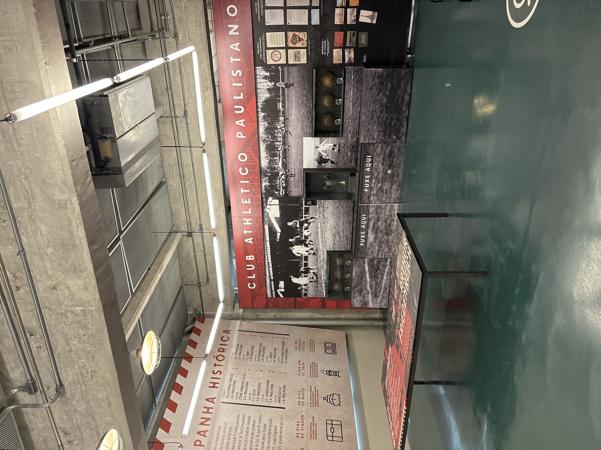 The image shows an exhibit dedicated to Club Athletico Paulistano in São Paulo, Brazil. A red and white banner with the club's name vertically displayed and a large section displaying historical facts, photos, and memorabilia dominate the scene. The exhibit includes a display titled "PANHA HISTORICA" with details of the club's history. A table with a sign encourages interaction. The setting appears to be a modern, industrial-style space.