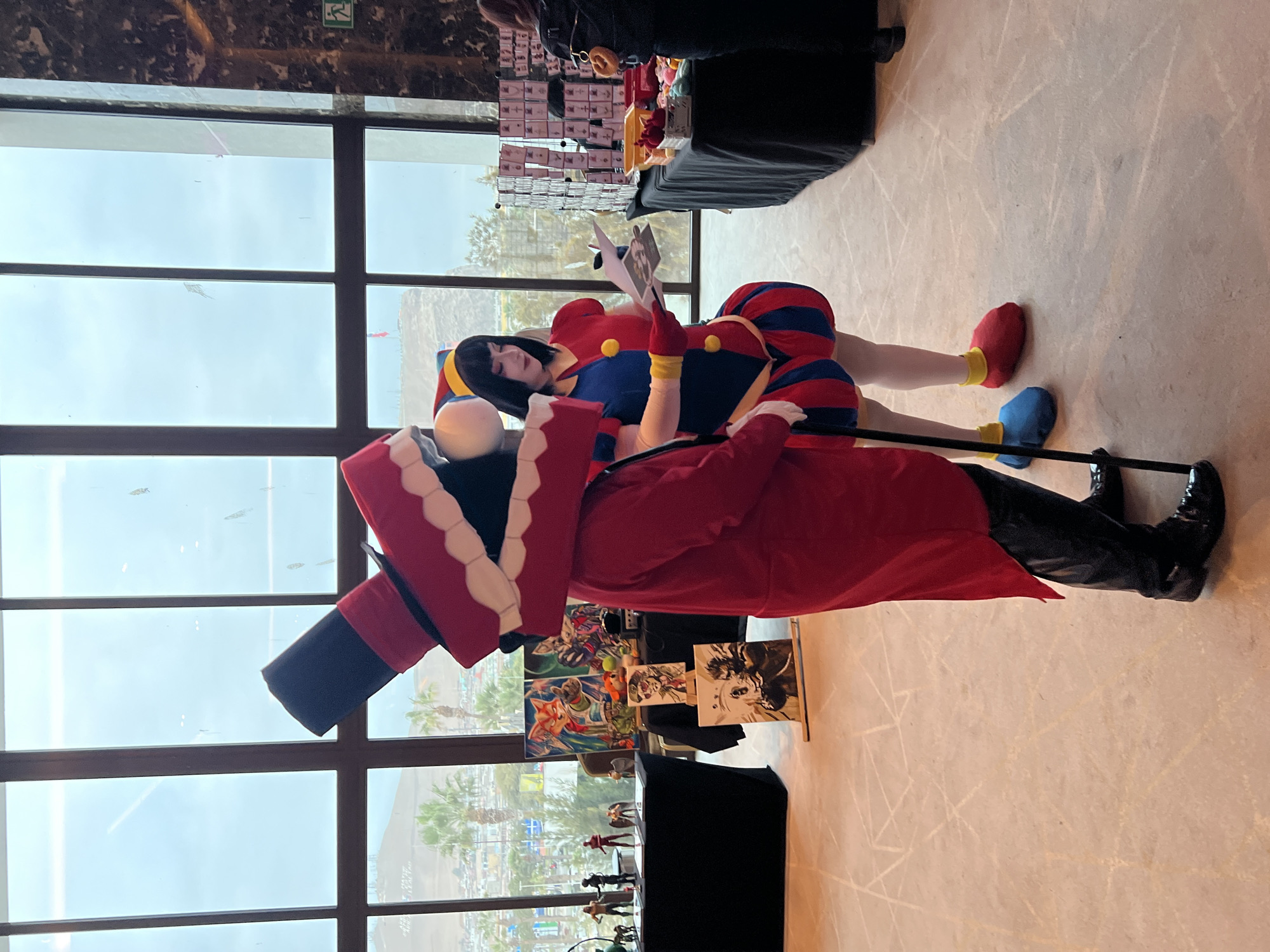 Two individuals in costumes stand in a brightly lit indoor space, likely a convention center, near large windows overlooking a city and ocean. The person on the left wears a large, red, cartoonish mouth mask over their head, a tall black top hat with a red band, and a long red coat with black pants and black dress shoes. They hold a black cane. The person on the right is dressed as a jester with a blue and red striped skirt, a yellow and red bodice with yellow pom-poms, a blue and yellow jester's hat, and red and blue shoe covers. They are looking down at a brochure or paper they are holding.
In the background, several tables are set up. One table on the left displays several small action figures and other merchandise. Further back, another table holds various artworks, including paintings of cartoon characters and possibly anime figures. A rack displaying various items, potentially jewelry or accessories, is visible on a table to the right. The floor has a light-colored, patterned carpet. The windows reveal a hazy view of buildings, palm trees, and a body of water, suggesting a coastal location. The presence of merchandise and costumes indicates an event like a comic convention or fan gathering. The architecture and landscape visible through the windows are consistent with the city of Arica, Chile, known for its coastal location and arid surroundings.