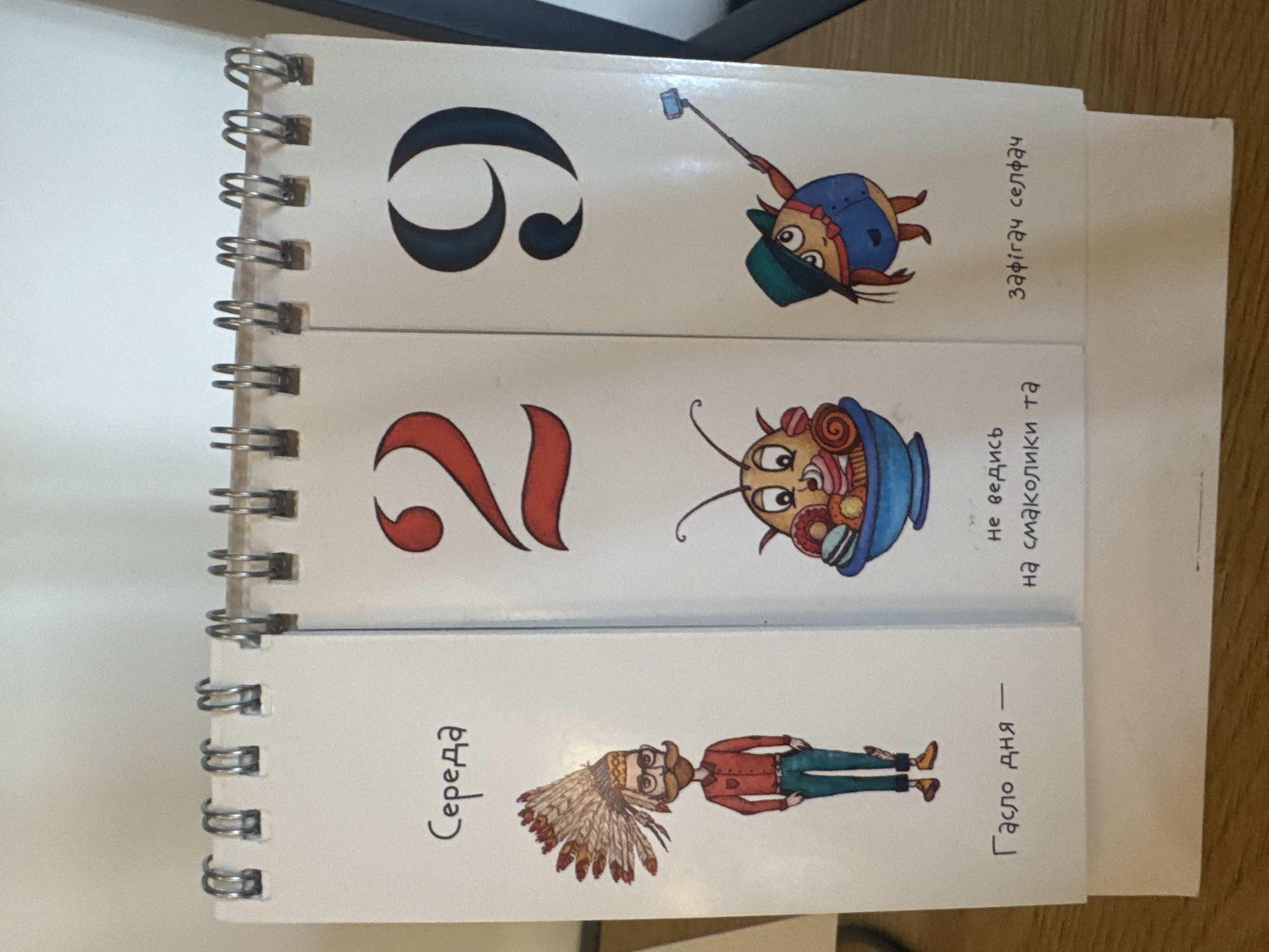 The image shows a desk calendar on a wooden surface. The calendar has three vertical panels. The left panel has the word "Середа" (Wednesday) written at the top, followed by a drawing of a man wearing a headdress, glasses, and a red shirt, with the text "Гасло дня -" (Motto of the day -) below. The middle panel displays the number "29" in red and a drawing of a character in a bowl of sweets, with the text "не ведись на смаколики та" (don't fall for sweets and) below. The right panel also displays the number "29" in dark teal and a drawing of a character taking a selfie with a selfie stick, with the text "зафігач селфач" (take a selfie) below. The calendar is bound with silver spiral wire. There are no people visible in the image, and the environment is a neutral indoor setting. The image does not provide any information about the location in the city of Kremenchuk, Ukraine.