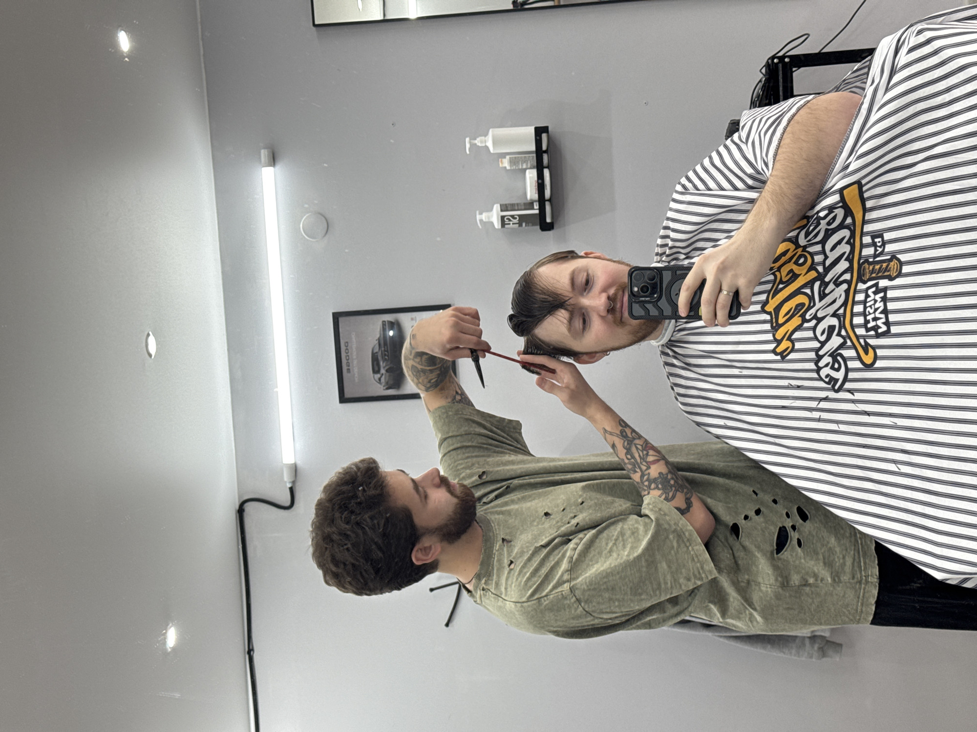 The image depicts an interior scene within a barbershop in Votkinsk, Russia. A male barber, with curly dark hair, a beard, and visible tattoos on his forearms, is positioned on the left, wearing a distressed olive-green t-shirt. He holds shears in his right hand and a comb in his left, actively cutting the hair of a male client seated to his right. The client has light brown hair, a light beard, and a nose stud, and is covered by a white and black striped barber's cape featuring a yellow and white logo. He is holding a black smartphone with multiple rear cameras in his right hand, directing it towards himself, suggesting he is taking a selfie or recording. The background consists of light grey walls. On the left wall, a black-framed picture displays a dark classic muscle car, with the word "DODGE" visible. To the right of the barber's head, a black rectangular shelf holds several white pump bottles and containers. A tall, narrow mirror is partially visible on the far right. Overhead, a long fluorescent tube light and recessed spot lights illuminate the area, and a white circular ventilation cover is present on the wall.