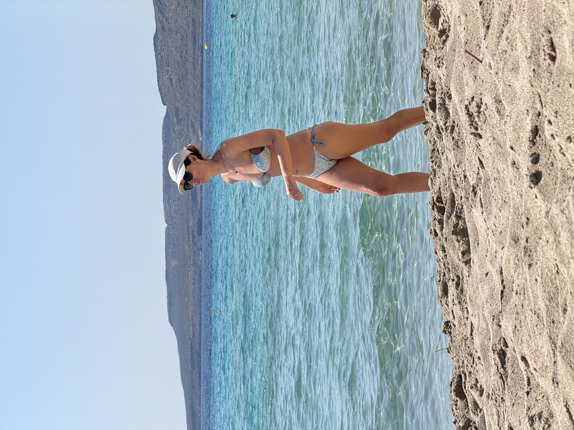 A woman in a white baseball cap and sunglasses, wearing a light blue patterned bikini, walks on a sandy beach towards the viewer. The water is clear and turquoise, with small waves. In the background, a hazy, dark green hill rises from the ocean under a clear blue sky. The sand in the foreground is light brown and textured.