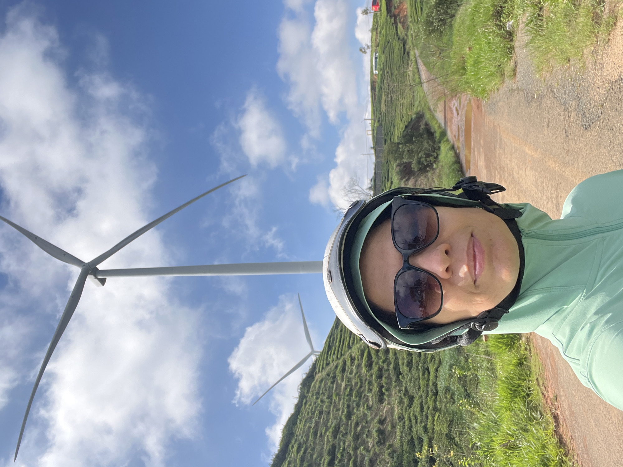 A woman wearing a green hooded jacket and a helmet with sunglasses is taking a selfie. She is positioned in the foreground, with a dirt road and lush green hills in the background. Two large wind turbines are visible on the hillside, with one closer to the camera and another further away. The sky is blue with scattered white clouds. The landscape appears to be a rural area, possibly a tea plantation, given the terraced greenery. The overall scene suggests an outdoor activity in a scenic location. Based on the context provided, the location is identified as Phát Chi, Vietnam.