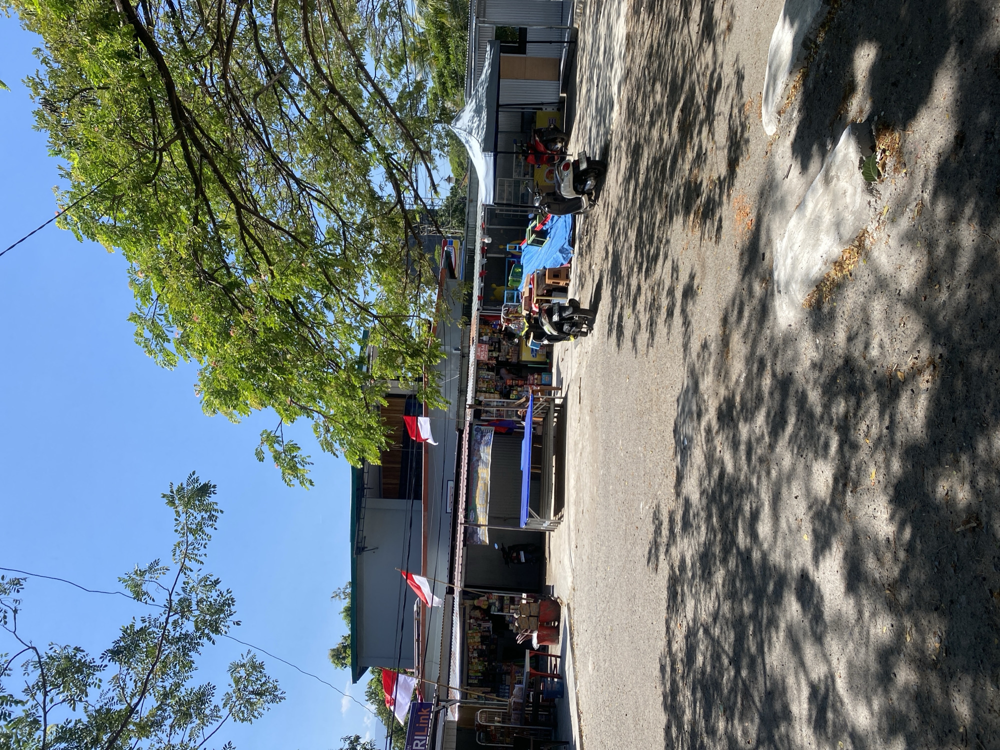 The image depicts a street scene in Bantaeng, Indonesia, likely in a commercial area. In the foreground, a paved road with dappled shade from overhead trees is visible, with some concrete blocks at the edge. A row of shops or stalls lines the background, featuring open fronts displaying various goods. Indonesian flags are prominently displayed, hanging from poles above the shops. Several motorcycles are parked and in motion. A blue tarp covers a section in front of one of the stalls, and a grey tent-like structure is also present. The environment appears to be outdoors on a bright, sunny day, with lush green trees framing the upper part of the image and a clear blue sky above.