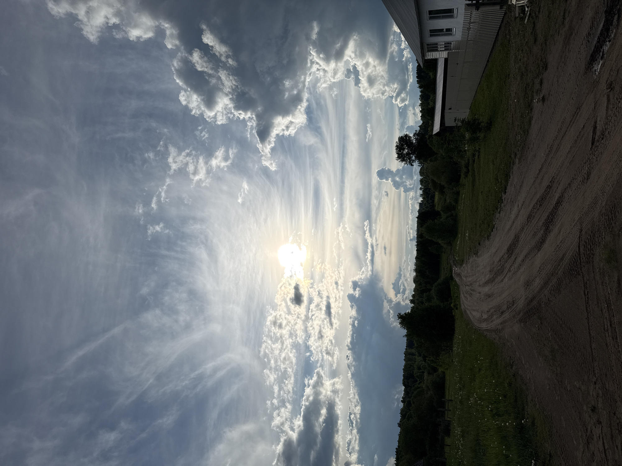 The image presents a daytime scene in what appears to be a rural or semi-rural setting. The sky dominates the view, filled with a mixture of cirrus and cumulus clouds, with the sun shining brightly through a break in the cloud cover. The landscape transitions to a dirt road on the right side of the image, exhibiting tire tracks. Adjacent to the road is a grassy area with wildflowers. A line of trees extends along the horizon. A building with white siding can be partially seen at the top right corner. The specific location is Уральское, Russia.