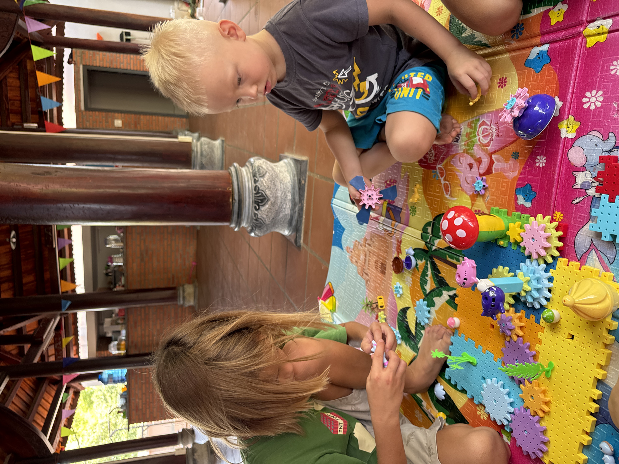 Two young children are captured in a bright, daytime setting, deeply engaged in playing with colorful interlocking gear toys on a vibrant playmat. The scene appears to be a semi-outdoor or open-plan indoor area, featuring architectural elements typical of Mũi Né, Vietnam.
A blonde-haired boy, positioned towards the upper right of the frame, wears a gray t-shirt with indistinct graphics and blue shorts with the visible text "RIDER UNIT". He is focused intently on a small pink gear in his hand, observing the toy setup on the mat. To the lower left, another child with light brown hair, wearing a green t-shirt with "TNT" printed on it, is also engrossed in manipulating the gear pieces.
The playmat beneath them is brightly colored in sections of pink, orange, yellow, light blue, and green, decorated with cartoon animals like elephants and stars. A variety of gear toys, including cogs and pieces shaped like a red mushroom, pink pig, and purple snail, are spread across the mat, some already interlocked to form a larger mechanism.
In the background, sturdy wooden pillars with ornate, grey bases are prominent, contributing to the traditional Vietnamese architectural aesthetic. Colorful triangular bunting in shades of red, blue, orange, green, and purple is strung across these wooden structures, adding a festive touch. Glimpses of a red brick wall and outdoor greenery suggest an open, airy environment. Both children appear calm and concentrated on their creative play.