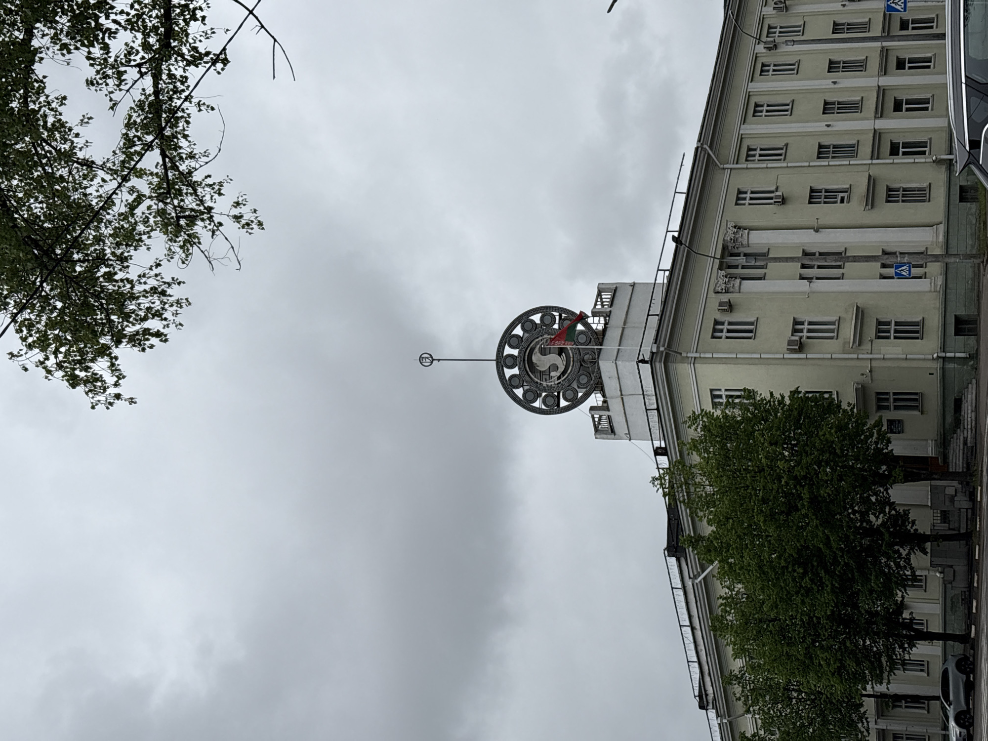 The image is a low-angle shot of a building in Minsk, Belarus, taken on a cloudy day. The building is several stories tall, light beige, and has numerous windows with a repeated pattern. A large decorative structure adorns the roof. It features a circular emblem with an animal or bird within it and a red flag. This emblem is set atop a white tower-like structure. A tall pole extends upwards from the very top of the emblem. A tree with green leaves partially obscures the building on the right side. Branches of another tree are visible in the upper left corner. The sky is overcast and grey. Part of a vehicle is visible in the far-right side of the image. The orientation of the image appears to be rotated.