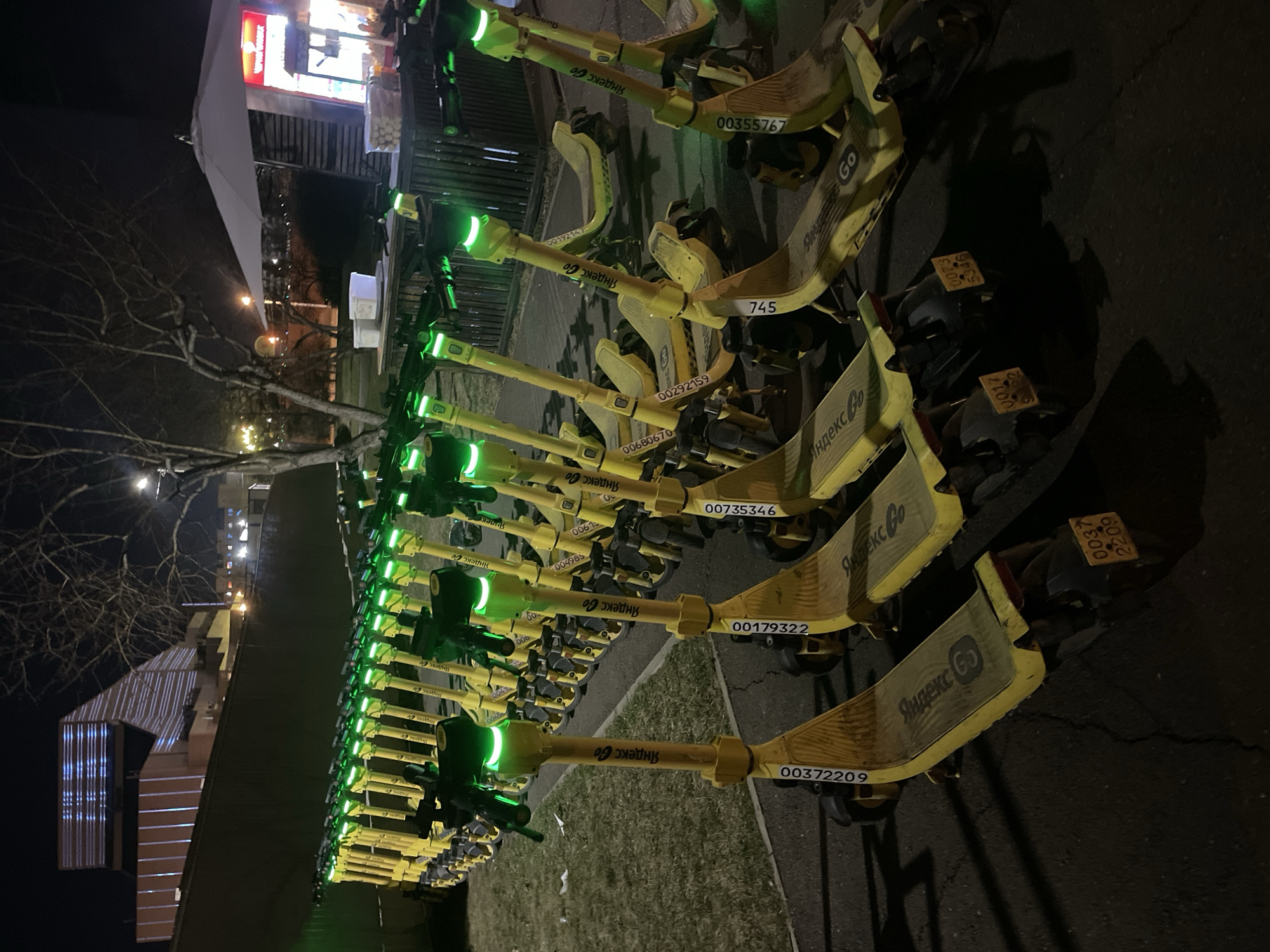 A fleet of yellow and black electric scooters, branded "Яндекс Go" (Yandex Go), is neatly lined up outdoors at night. Each scooter features prominent green glowing lights on its handlebars, likely indicating their status or charge. Many scooters have visible identification numbers, such as 00372209, 00179322, 00735346, 00292159, 00680670, 00355767, and 745. The setting is a paved area, possibly a sidewalk or parking zone, with a bare-branched tree suggesting a colder season like late autumn or winter. In the background, illuminated buildings, including what appears to be a commercial establishment with a red sign and an umbrella, indicate an urban environment. The scene is lit by artificial lights from the scooters themselves and distant sources, confirming it is nighttime. No people are visible. The scooters appear to be part of a shared mobility service in Krasnoyarsk, Russia, parked and awaiting users.