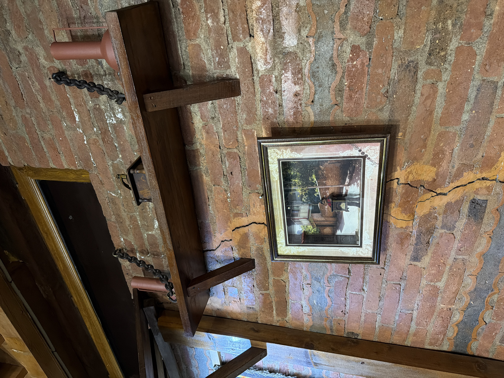 The image shows a close-up of a brick wall in what appears to be an interior setting in Tbilisi, Georgia. A wooden shelf is mounted on the wall, with a dark chain hanging from it. On the shelf, there are two decorative objects: a cylindrical brown object to the left and a red lamp-like object to the right, with another dark chain connected to it. A framed picture hangs on the brick wall below the shelf, depicting an outdoor scene with buildings and greenery. The brickwork itself shows signs of age and has some visible cracks. The overall impression is of a rustic or historic ambiance.