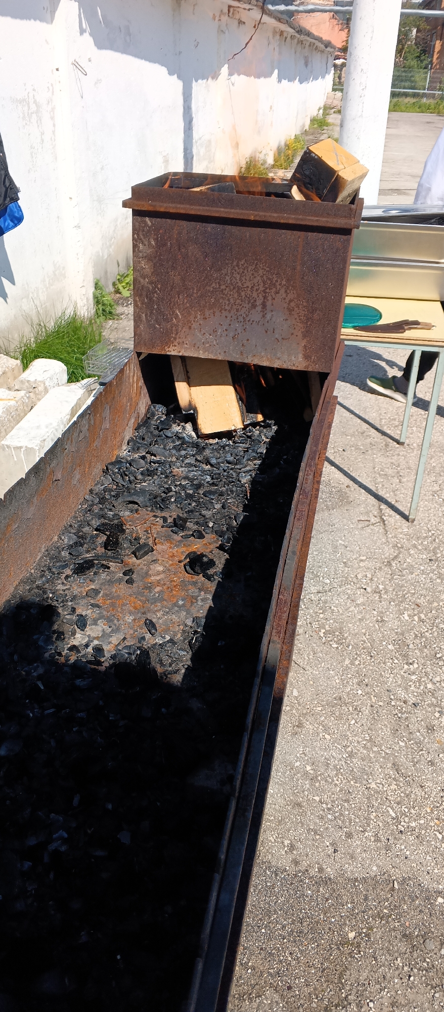 The image depicts an outdoor scene with a rusty, elongated metal barbecue pit filled with charcoal and burning wood. The pit is positioned on a gravel surface. To the left of the pit, there is a white concrete structure and some green grass. In the background, a white wall with shadows is visible. To the right of the barbecue, a metal table with a green circular object and knives is present, and a person's legs, wearing a green shoe, are visible. A white sheet or garment is also visible in the background on the right. The lighting suggests it is a sunny day, casting shadows.