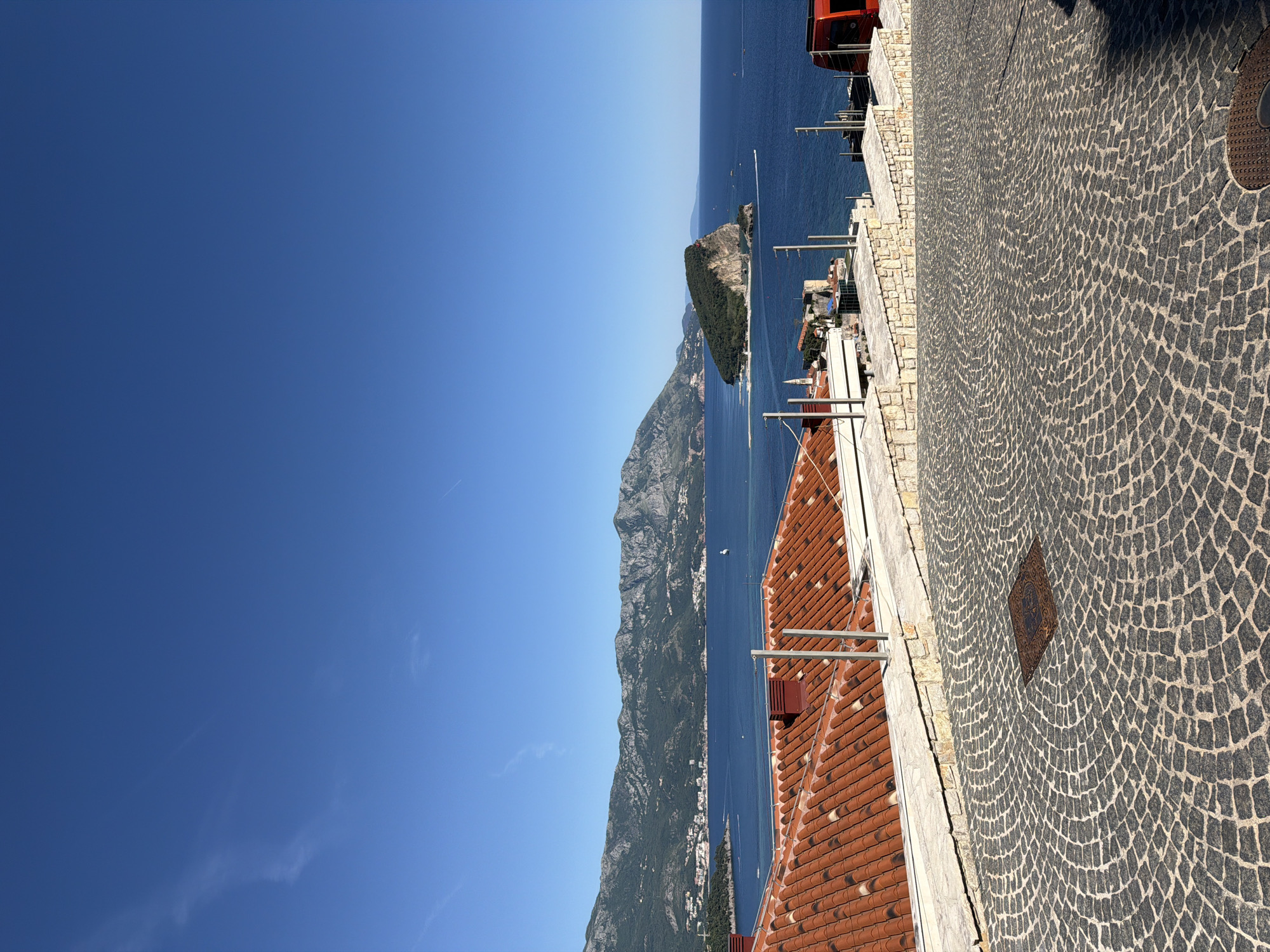 The image shows a view from a high point in Budva, Montenegro, looking out over the Adriatic Sea. The sky is clear and blue. In the distance, a rocky island and the coastline are visible. A stone-paved street occupies the foreground, with terracotta rooftops of buildings slightly below the viewpoint. The scene appears to be taken on a sunny day.