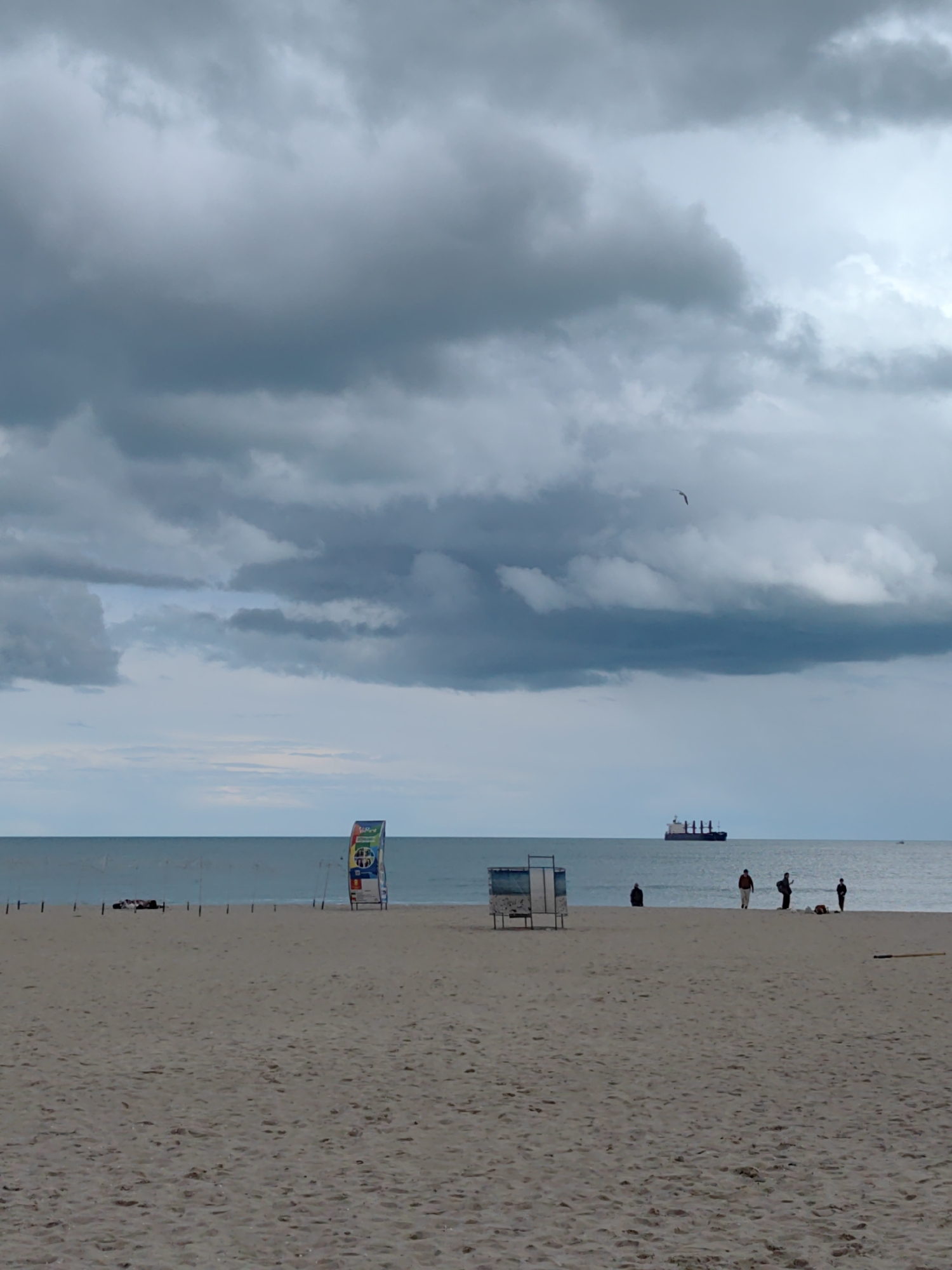 The image captures a cloudy day at a beach in Varna, Bulgaria. The sky is filled with heavy, gray clouds. The sand is light brown. Several people are on the beach: one person is sitting, and a group of three are standing. There is also a large cargo ship visible on the horizon. Beach furniture is visible, including a lifeguard stand and beach shelter.