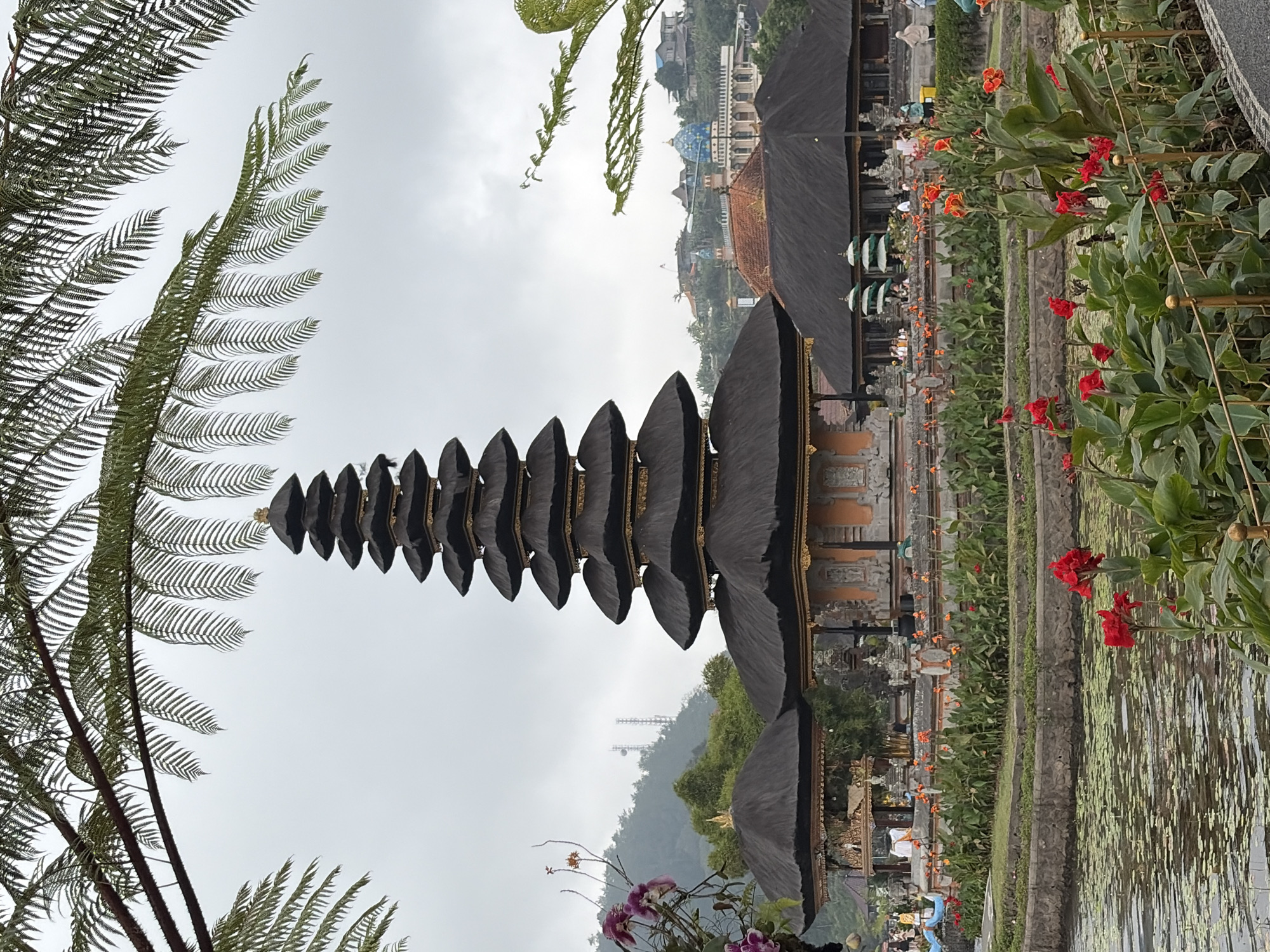 This is a vertical, full shot of the Pura Ulun Danu Beratan temple complex in Baturiti, Indonesia. The temple is situated on the edge of a body of water, which is partially covered with lily pads. In the foreground, bright red flowers are in bloom along a golden fence. Behind the water and flowers, a large, multi-tiered temple structure with a dark thatched roof and ornate gold trim is the focal point. Several smaller structures with similar thatched roofs are also visible. People can be seen walking around the temple grounds. In the background, lush green hills rise, dotted with buildings and two radio towers. The sky is overcast and a large fern frond hangs over the scene from the top left, with another smaller frond to the top right.