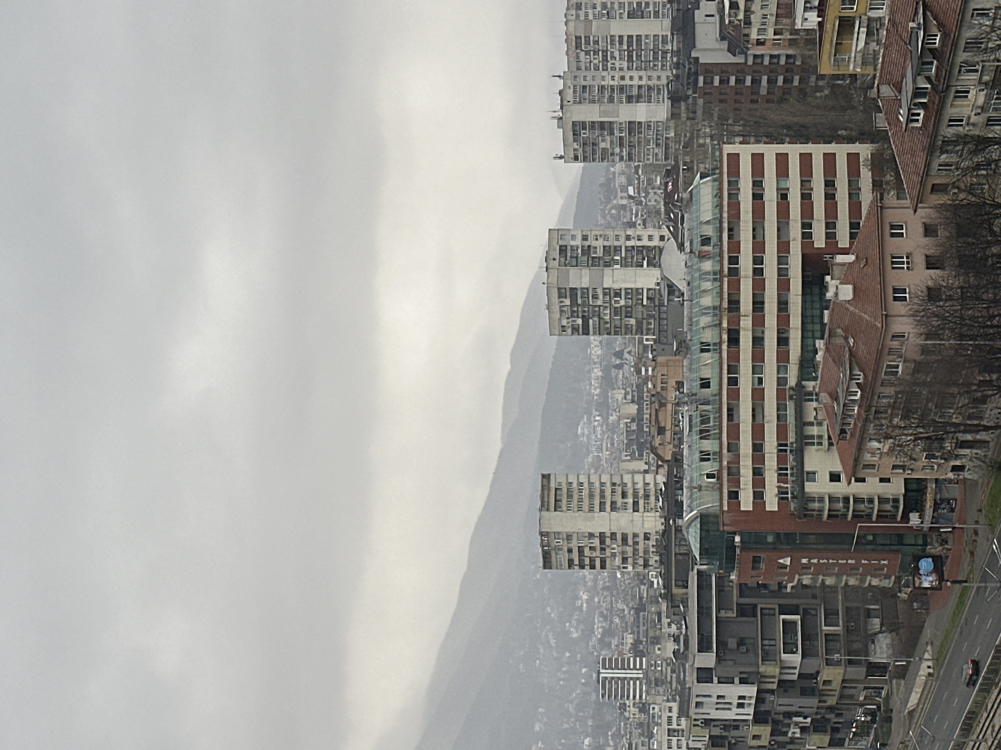 An elevated, wide-angle view captures a segment of the city of Sofia, Bulgaria, under an overcast sky. The scene is dominated by urban architecture with the Vitosha mountain range faintly visible in the distant background, partially obscured by atmospheric haze.
In the foreground, a multi-lane asphalt road runs diagonally from left to right, bordered by a sidewalk and bare deciduous trees. A single dark-colored sedan is visible on the road, traveling away from the viewer. Parallel to the road and to its left are railway tracks.
The midground features a dense collection of buildings exhibiting diverse architectural styles and ages. Prominently displayed is a multi-story building with a red-brick facade patterned with white horizontal stripes and a distinctive green, segmented glass dome roof structure. Adjacent to it, to the left, is a more contemporary, geometrically complex building constructed from varying shades of grey, white, and dark paneling. A vertical sign bearing the text "MASTER FIX" is visible on the side of a reddish-brown building located behind the road, near the geometric complex. Interspersed among these are older, smaller residential structures with reddish-brown tile roofs.
The background consists of numerous multi-story residential apartment blocks, primarily constructed of light-colored concrete, characterized by a utilitarian design, appearing to be older, potentially brutalist-style structures. These buildings are scattered across the urban landscape, extending towards the foothills of the mountains.
No individuals are clearly discernible within the frame. The overall atmosphere is muted, consistent with a cloudy day. The bare trees suggest either late autumn, winter, or early spring. This specific vantage point, including the visible "MASTER FIX" building and the railway lines with Vitosha mountain in the background, indicates a location within Sofia, likely viewed from an elevated position in the southwestern part of the city, possibly around the Bulgaria Boulevard area.