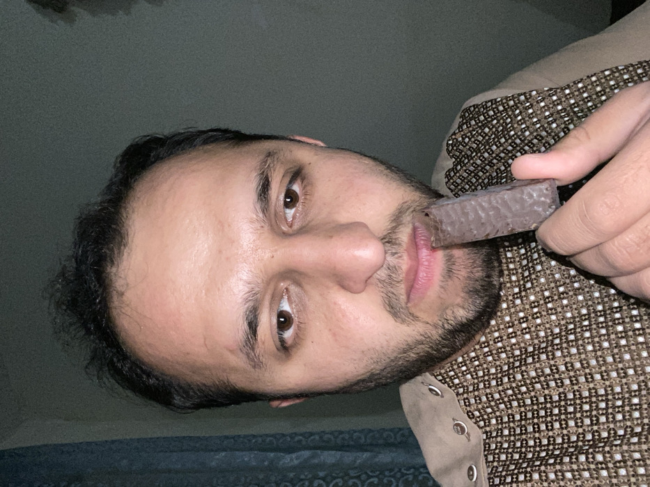 A close-up portrait shot depicts an adult male indoors, reportedly in Kandahar, Afghanistan, though no specific location within the city is discernible. The man has dark, slightly disheveled hair, a full beard, and a mustache. His eyes are dark brown, and he exhibits a medium skin tone with a minor blemish on his forehead. He wears a light-colored, long-sleeved garment, possibly a tunic, featuring a patterned brown and white checkered or dotted design across the chest, with visible snap fasteners along the left collarbone area. He holds a rectangular chocolate-covered wafer bar, which appears partially consumed, up to his mouth with his right hand; his lips are parted and in contact with the bar. His gaze is directed forward, slightly downward, towards the camera. The background consists of a dark blue or teal patterned curtain on the left and a plain, dark gray wall on the right. The lighting is dim, suggesting an indoor, low-light environment.